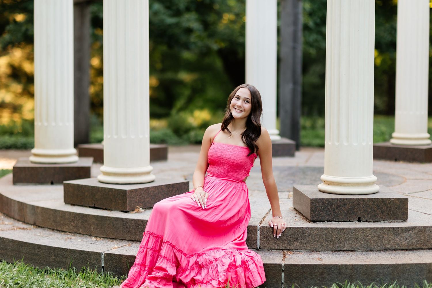 Young woman in pink maxi dress sitting on steps near white columns outdoors.