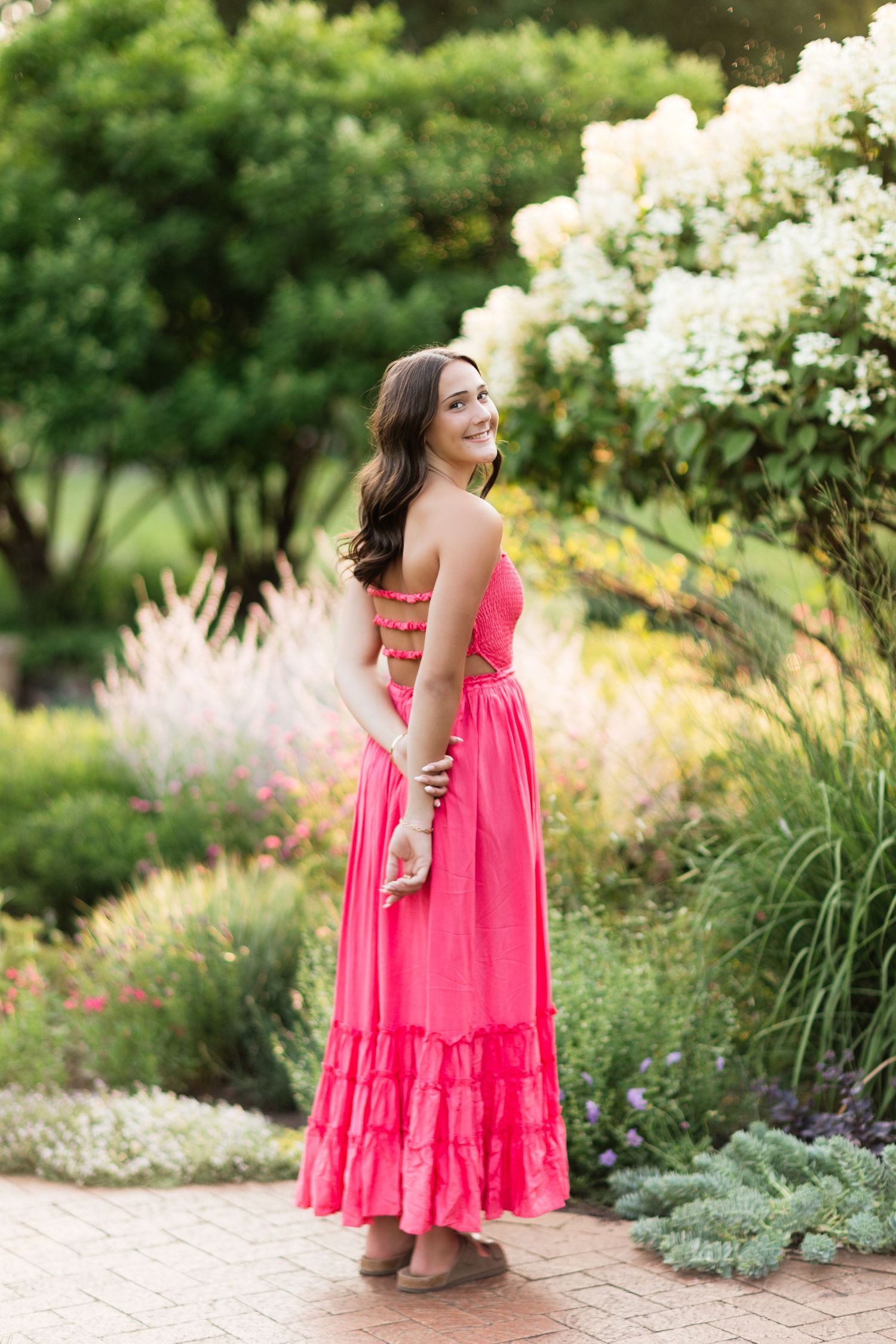 Young woman in pink maxi dress smiling in lush garden with white flowers.