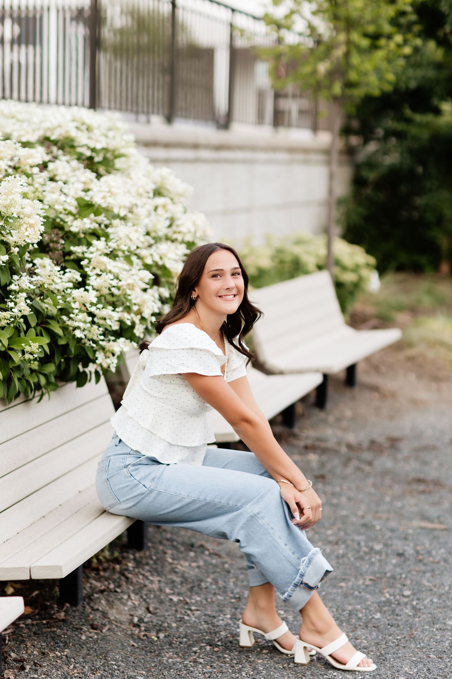 Young woman in white ruffled top and light jeans sitting on bench near white hydrangeas outdoors.