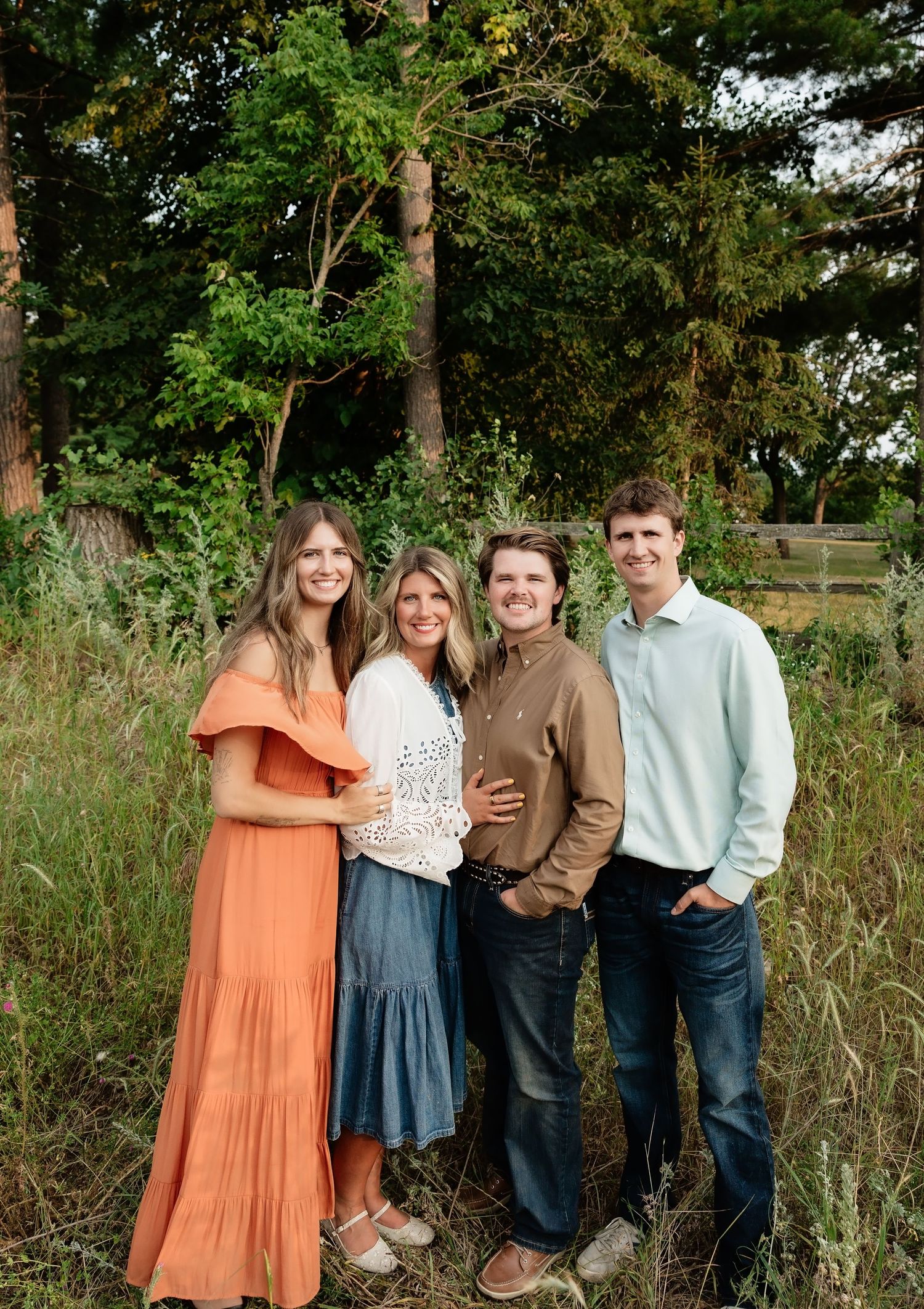 A joyful family of four poses outdoors in a lush green setting, wearing casual earth-tone outfits.