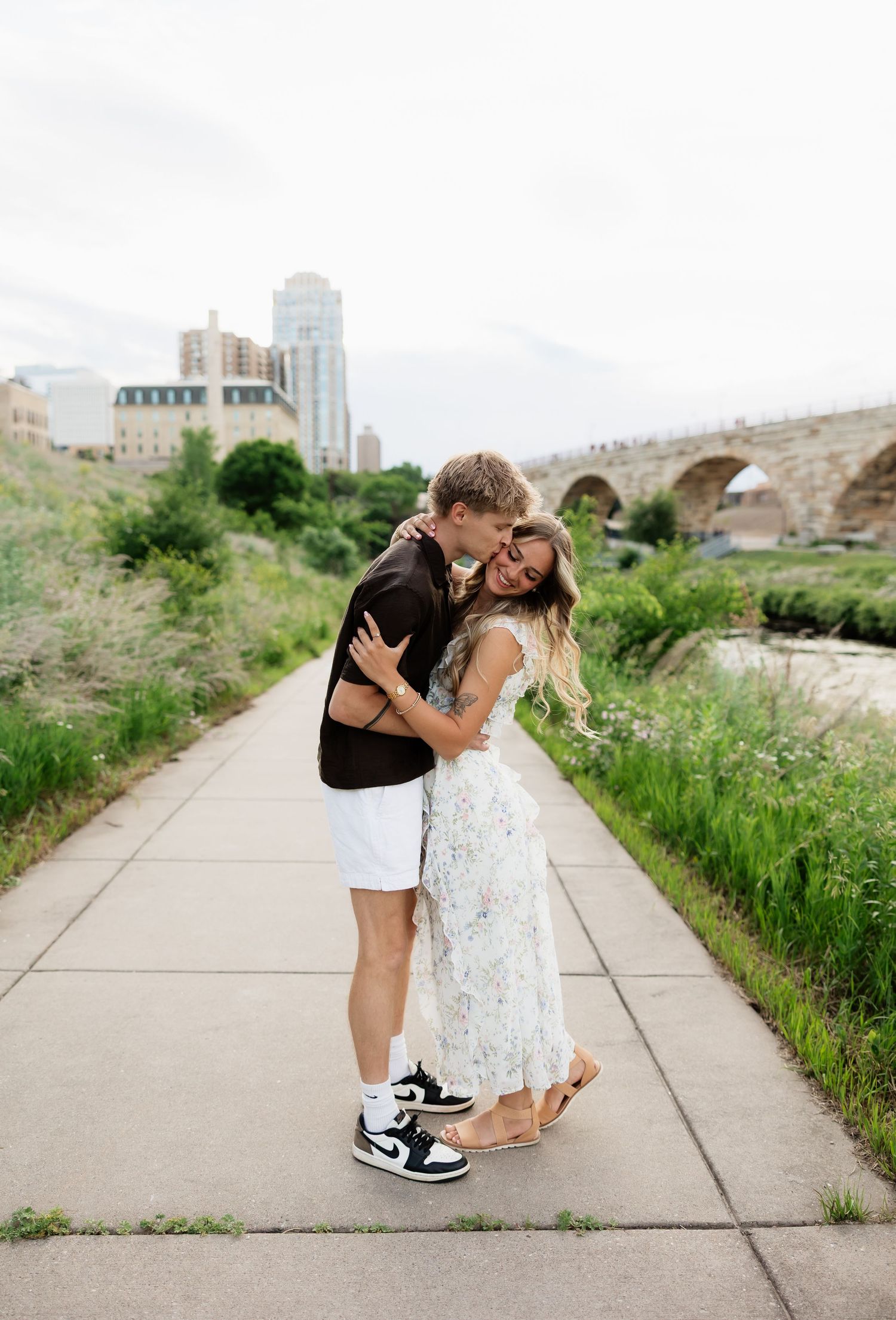 Romantic couple embracing and kissing on a riverside pathway near the Stone Arch Bridge in Minneapolis.