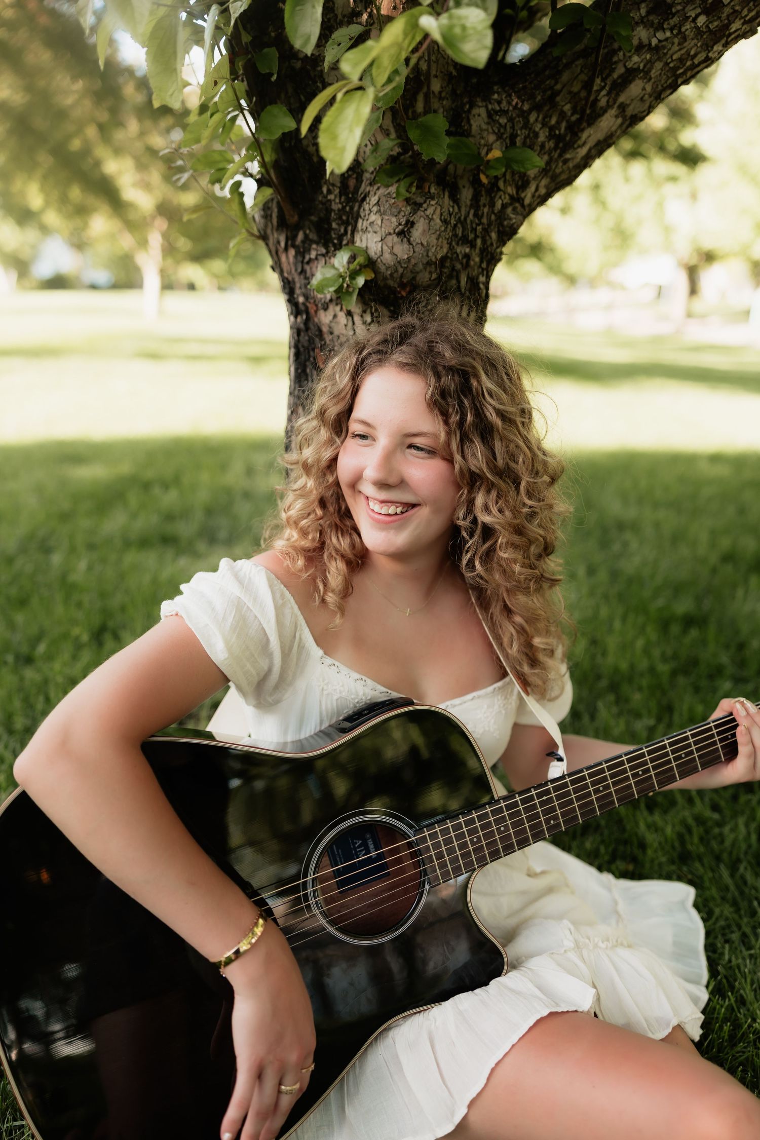 Smiling teenage girl in white dress holding an acoustic guitar while sitting on green grass under a tree.