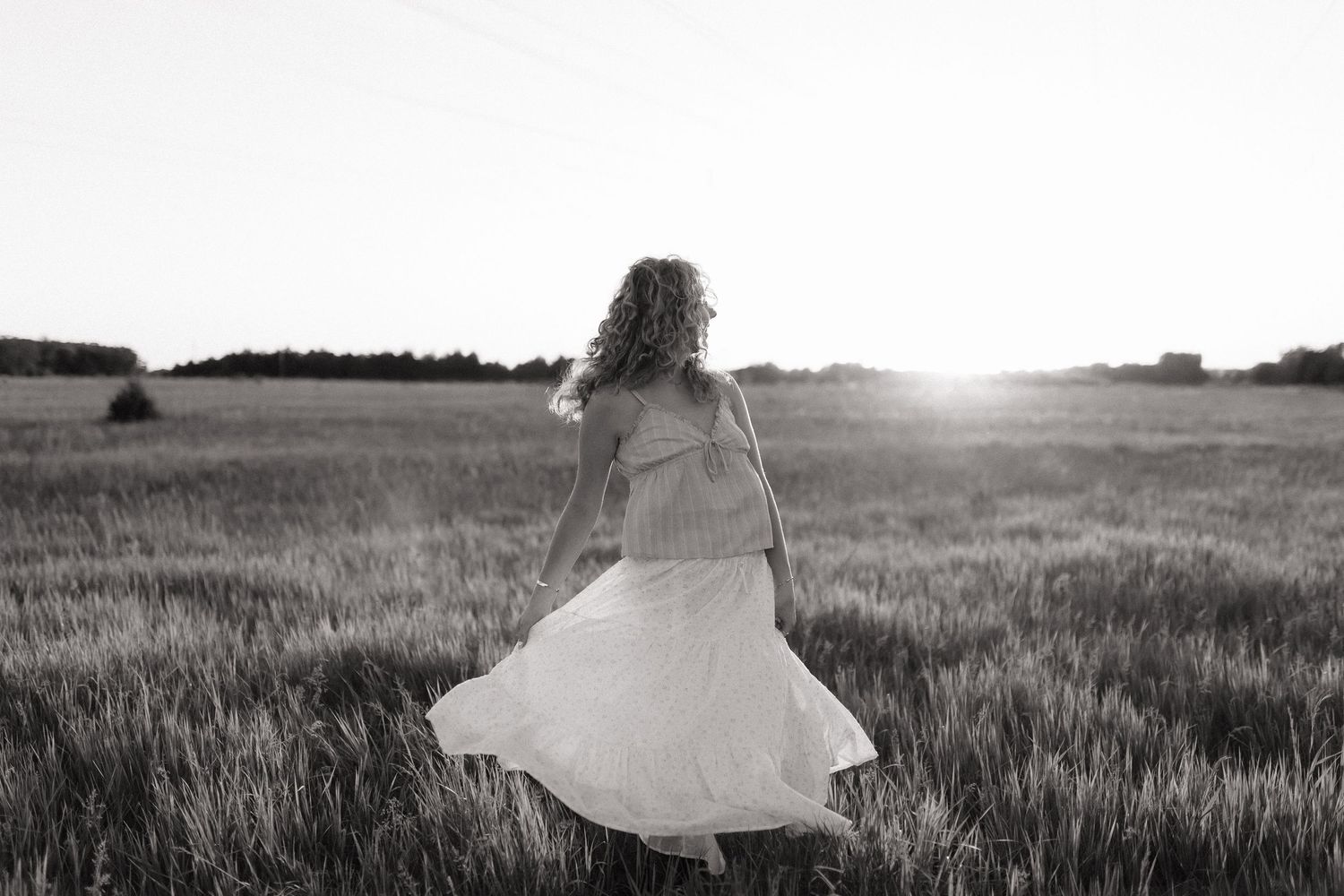 Black and white photo of woman in flowing white dress walking through field at sunset.