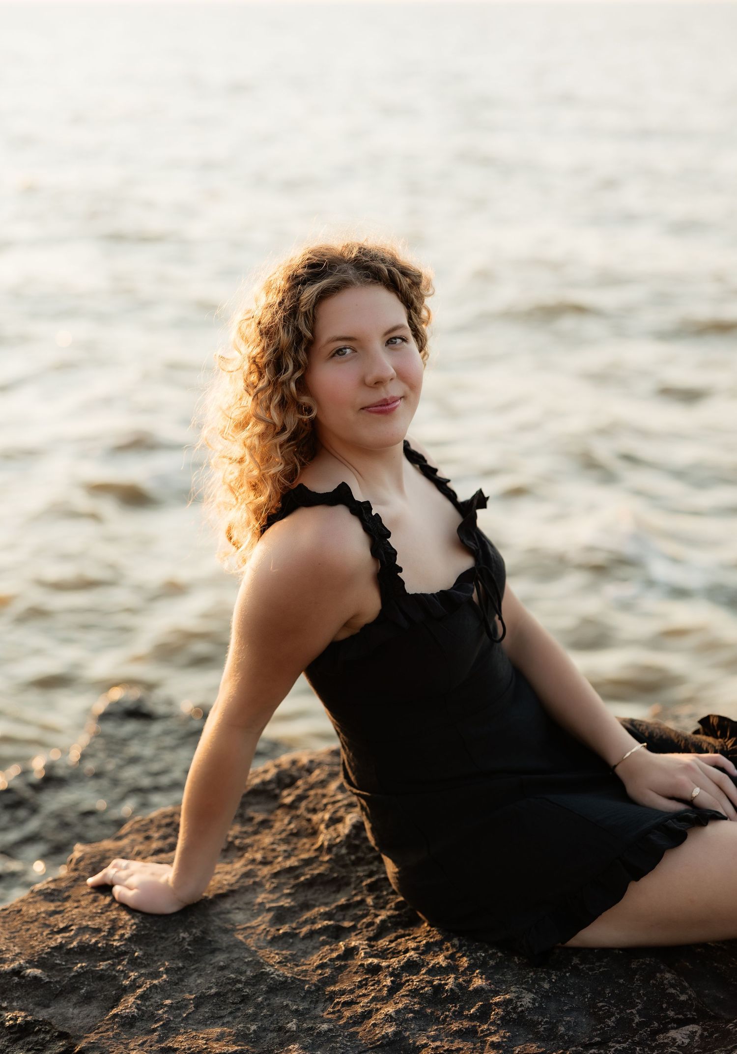 Curly-haired woman in black dress sitting on rocky shoreline with golden sunlit water behind her.