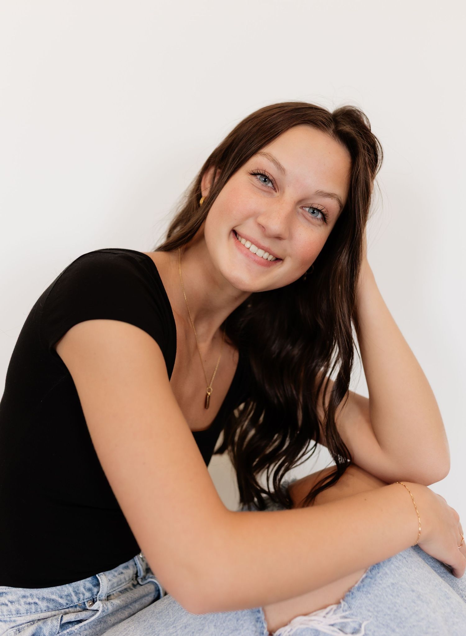 Smiling young woman with wavy brown hair in black tee and jeans posing against white background.