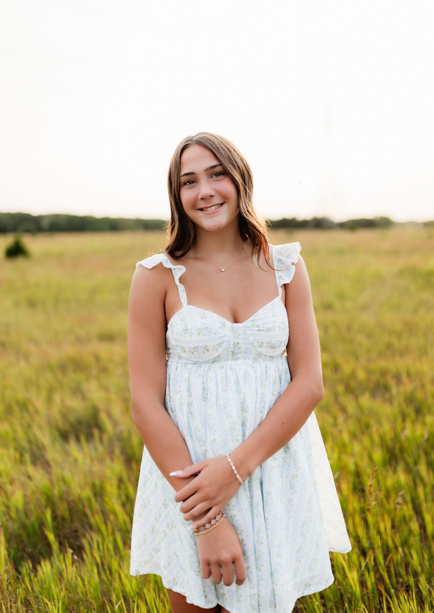 Smiling teenage girl in light blue sundress standing in open grassy field at golden hour.