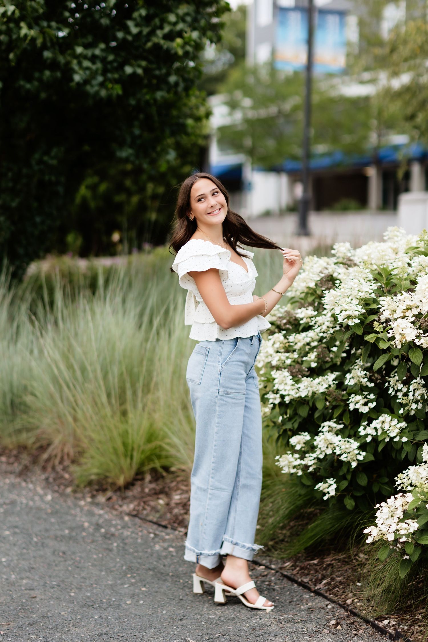 Happy young woman in white top and light jeans posing by white flowering shrubs on city pathway.