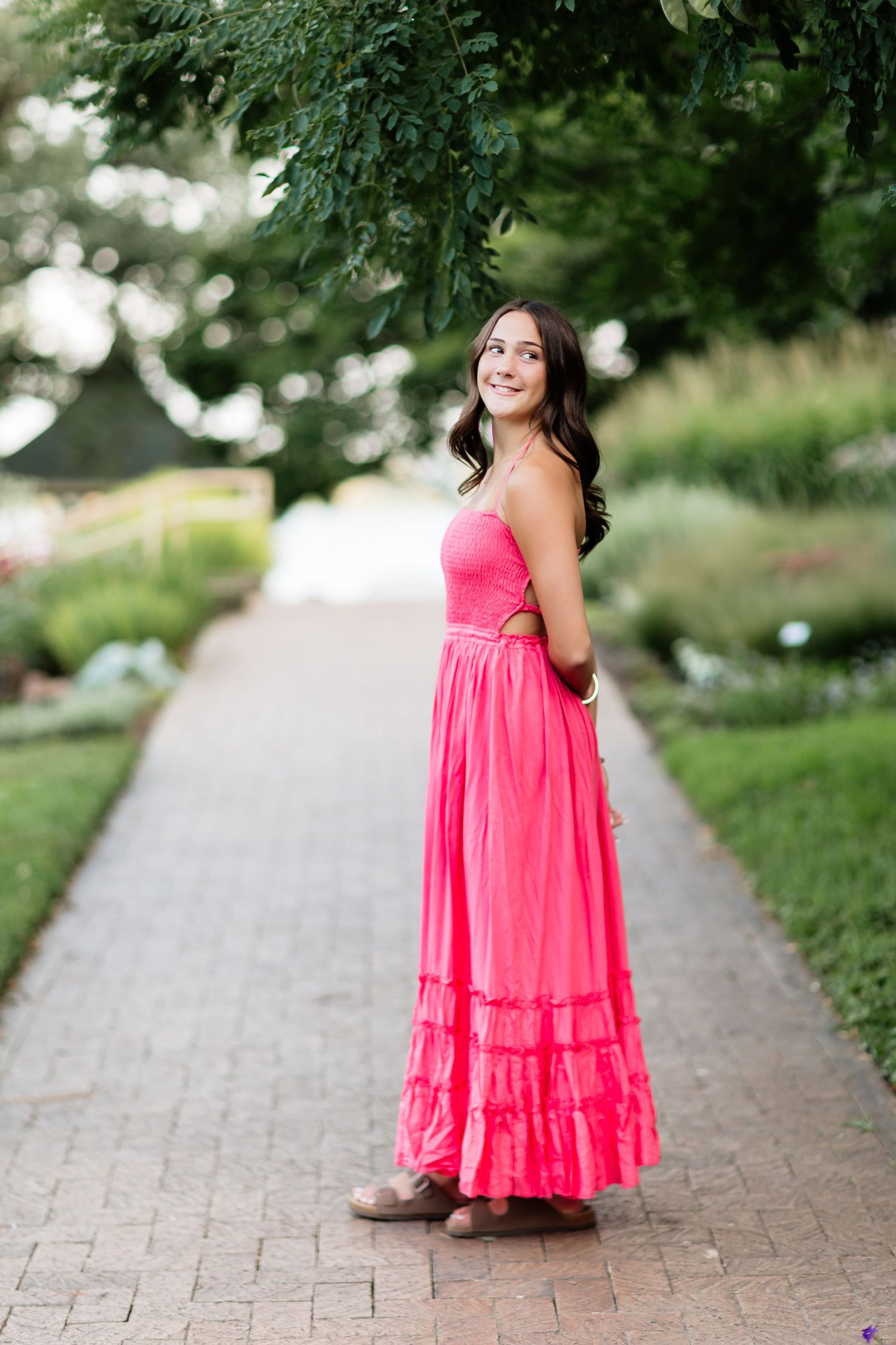 Young woman in a bright pink strapless maxi dress standing on a brick garden path under green trees.