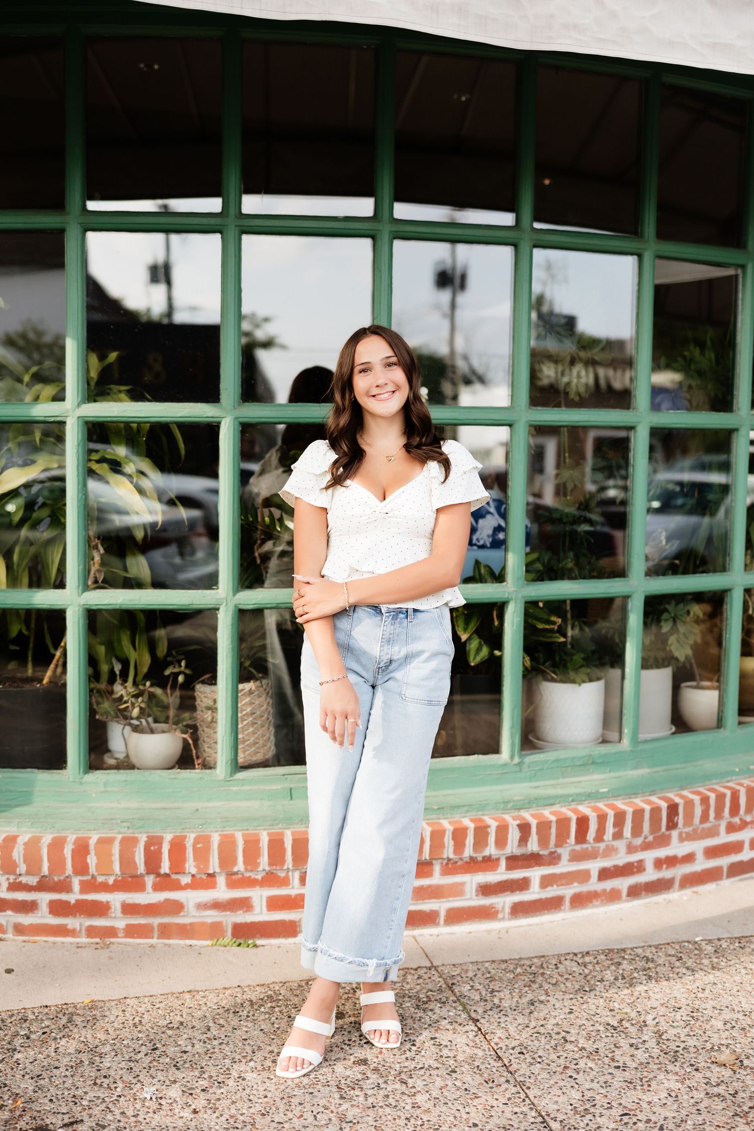 Young woman in white top and wide-leg jeans standing in front of a green-framed storefront.
