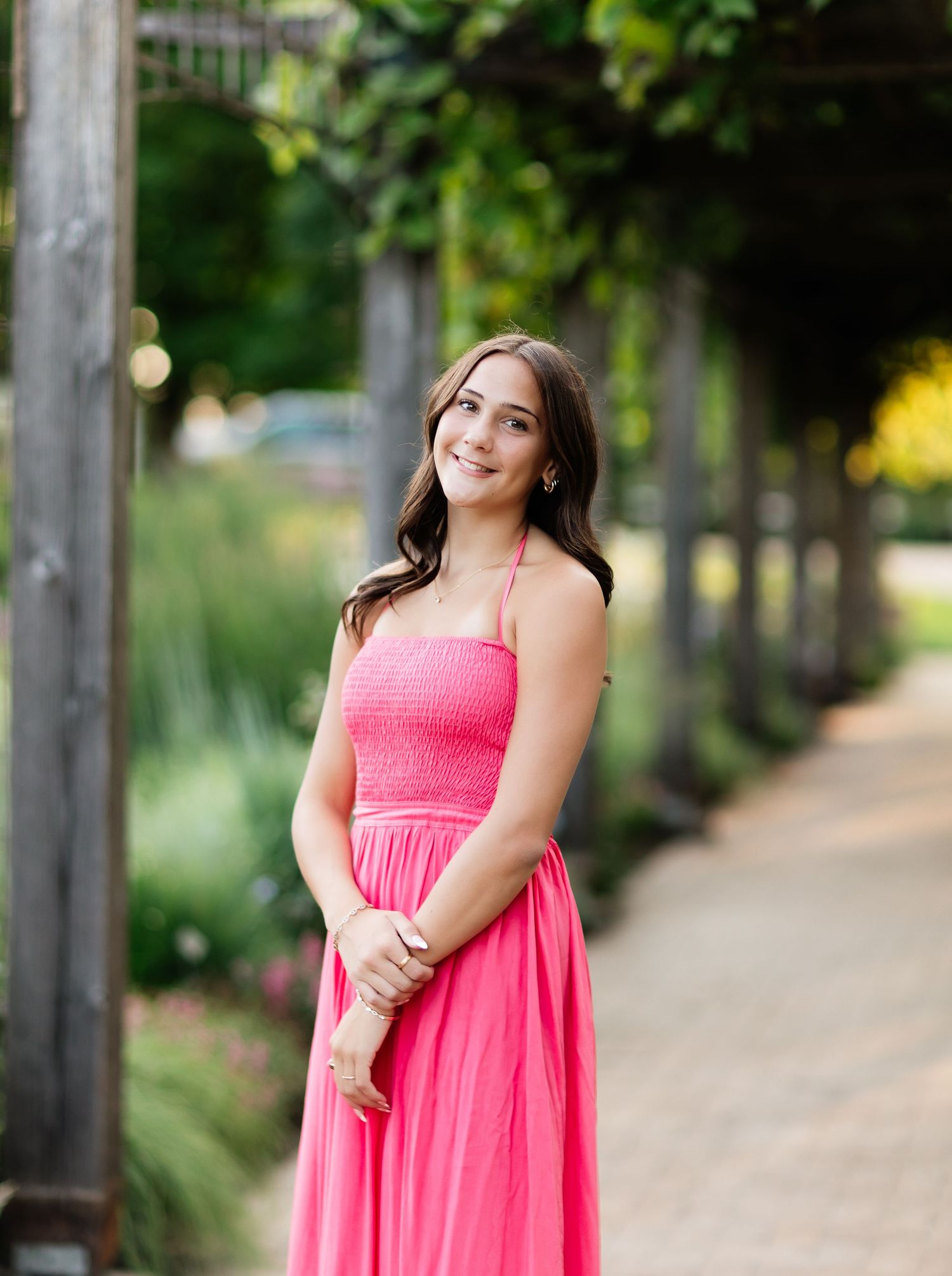 Young woman in pink maxi dress smiling on garden pathway with lush green pergola backdrop.