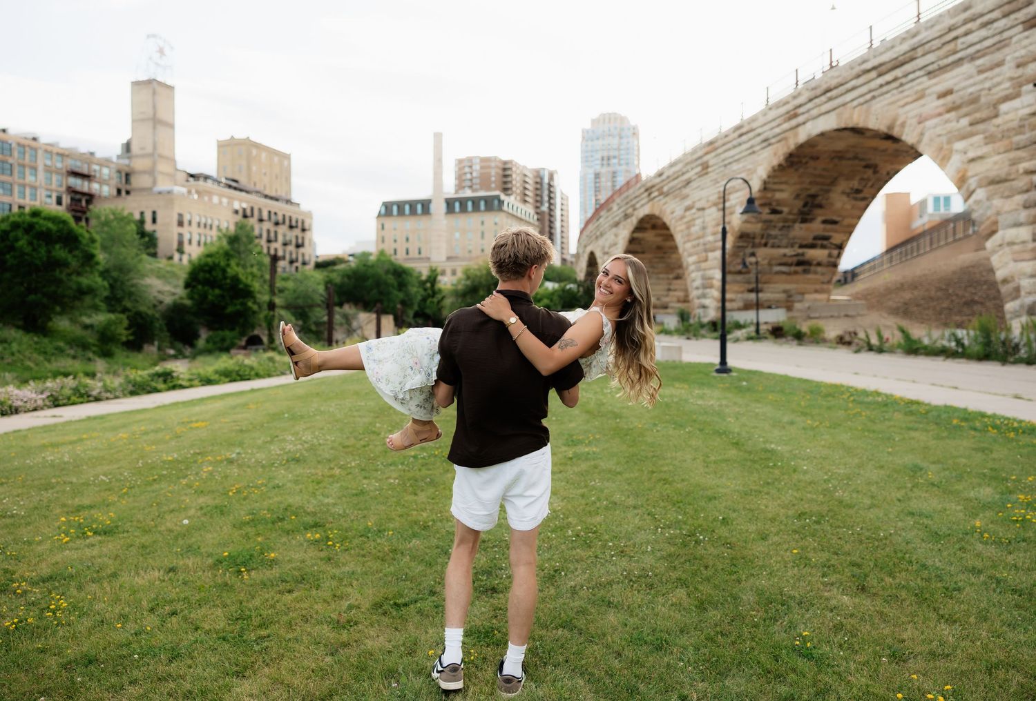 A man lifts and spins a woman in a floral dress on green grass with a historic stone arch bridge behind them.