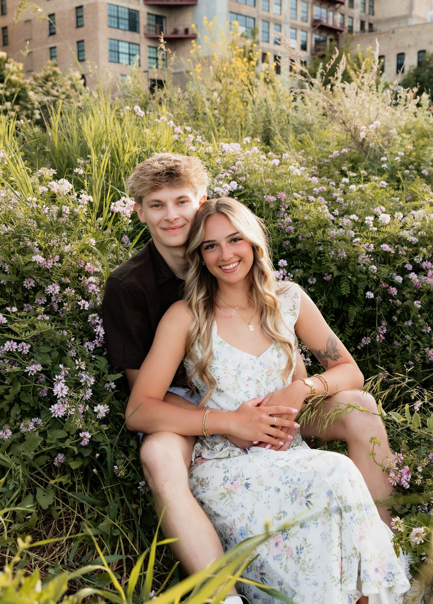 Couple sitting together in lush wildflower garden with brick buildings in soft golden background.