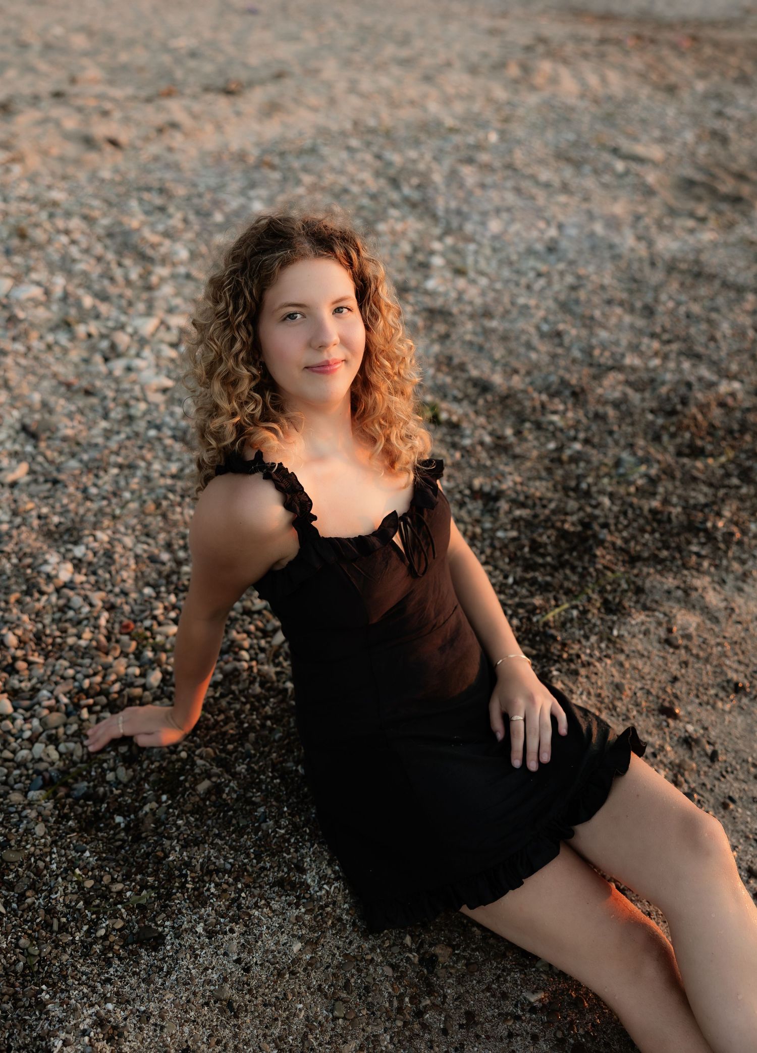 Young woman with curly auburn hair in black sundress posing on sandy beach at sunset.