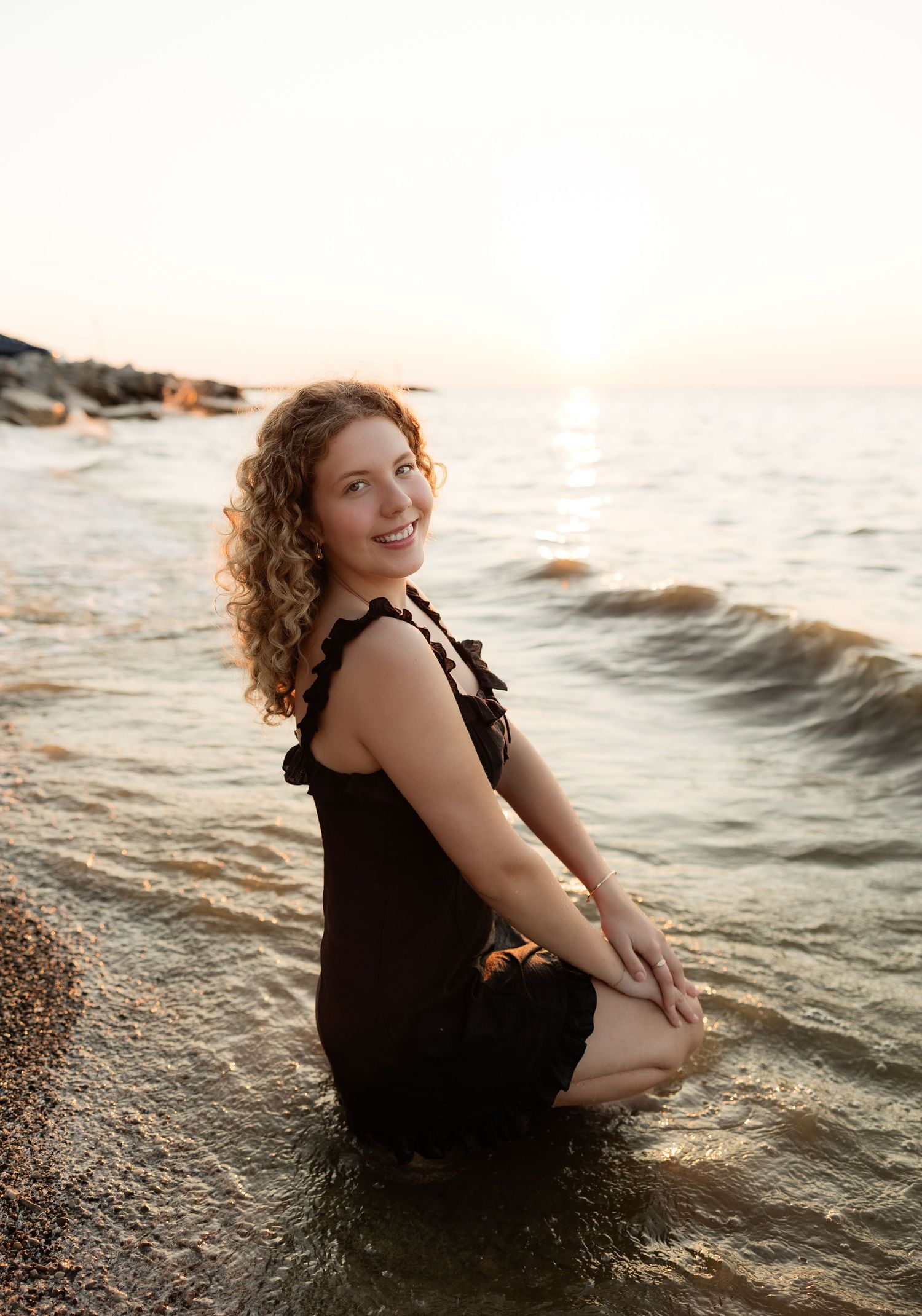 Young woman with curly hair smiling in black dress sitting in shallow ocean waves at sunset on rocky beach.