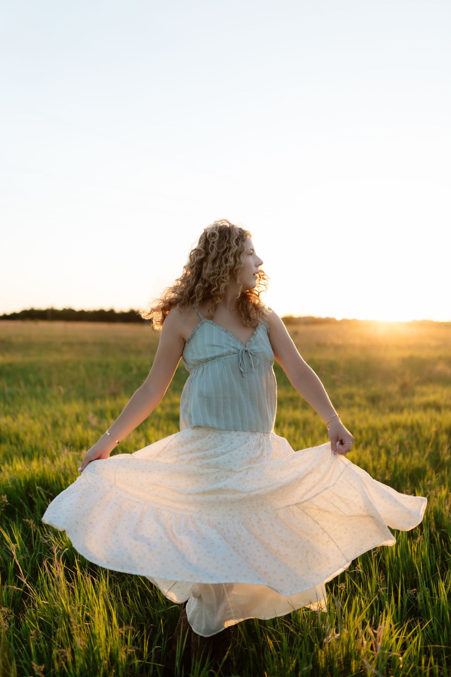 Young woman in white dress twirling in golden sunset field, curly hair flowing freely in warm summer light.