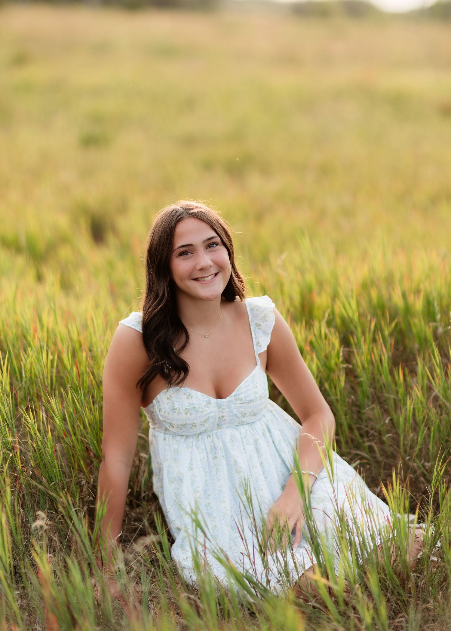 Brunette girl in white floral dress sitting relaxed in a lush green meadow, smiling warmly.