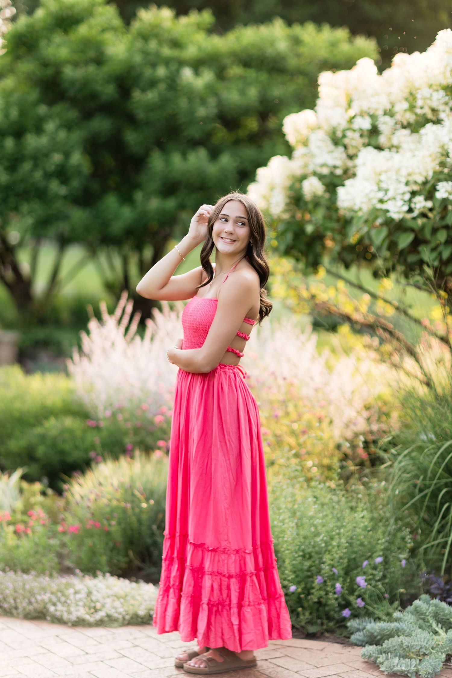 Young woman in bright pink maxi dress standing in lush garden with white hydrangeas blooming.