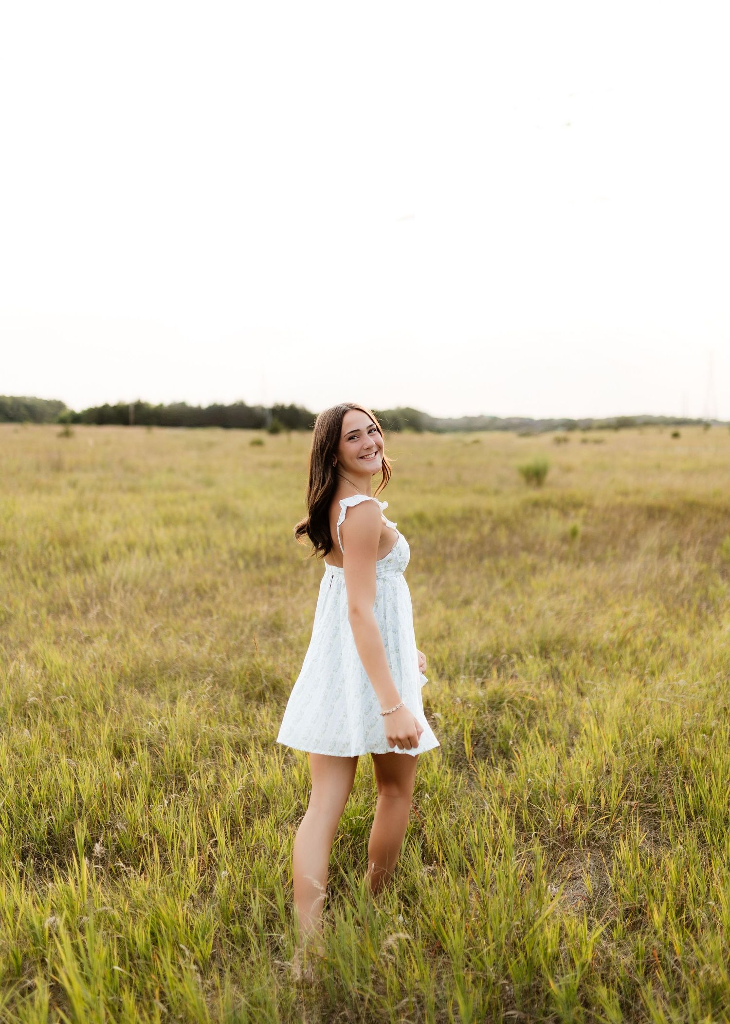Smiling young woman in white dress twirling in open grassy field at sunset.