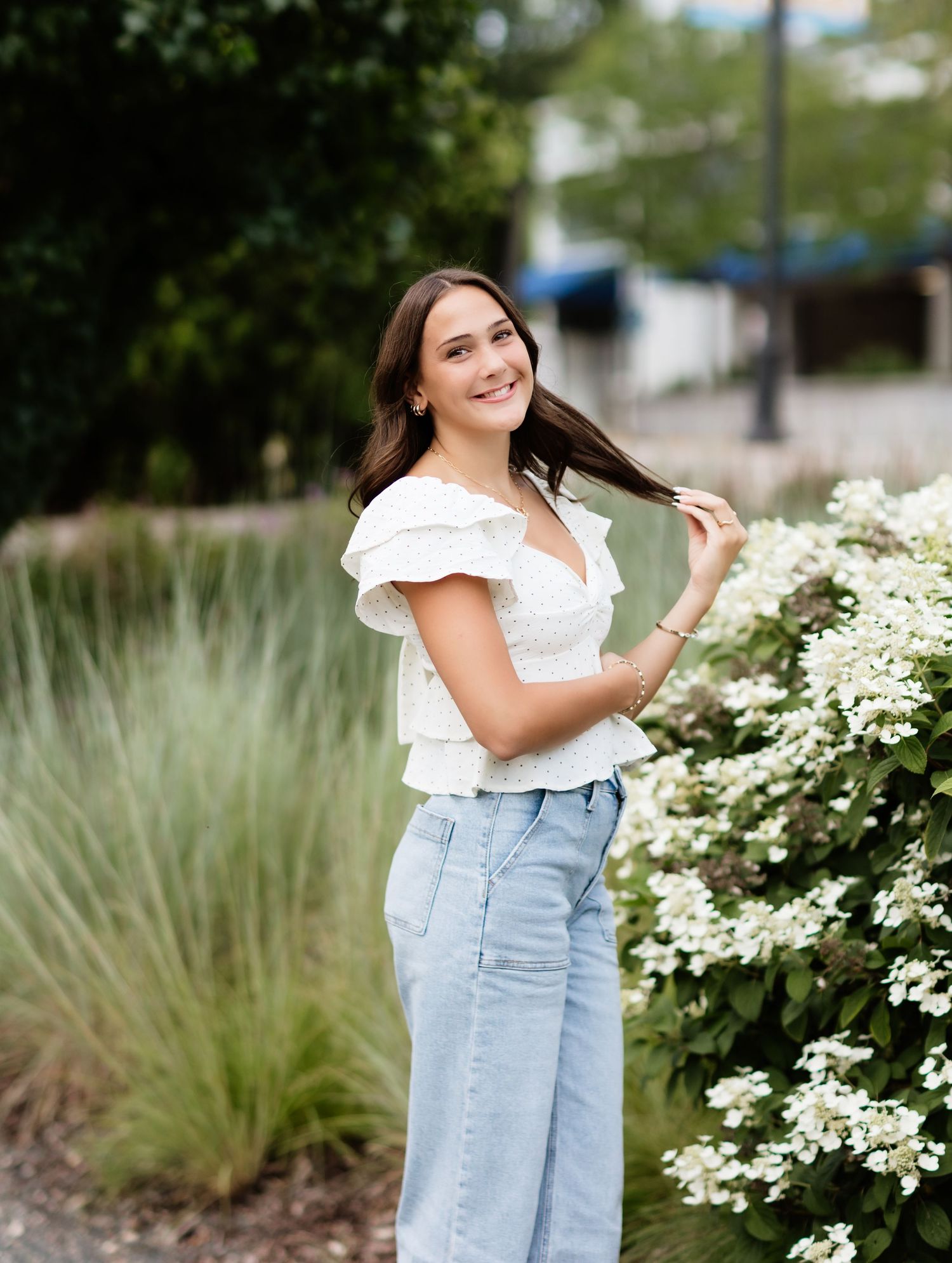 Young woman in white eyelet top and denim skirt laughing while touching her hair near white flowers.