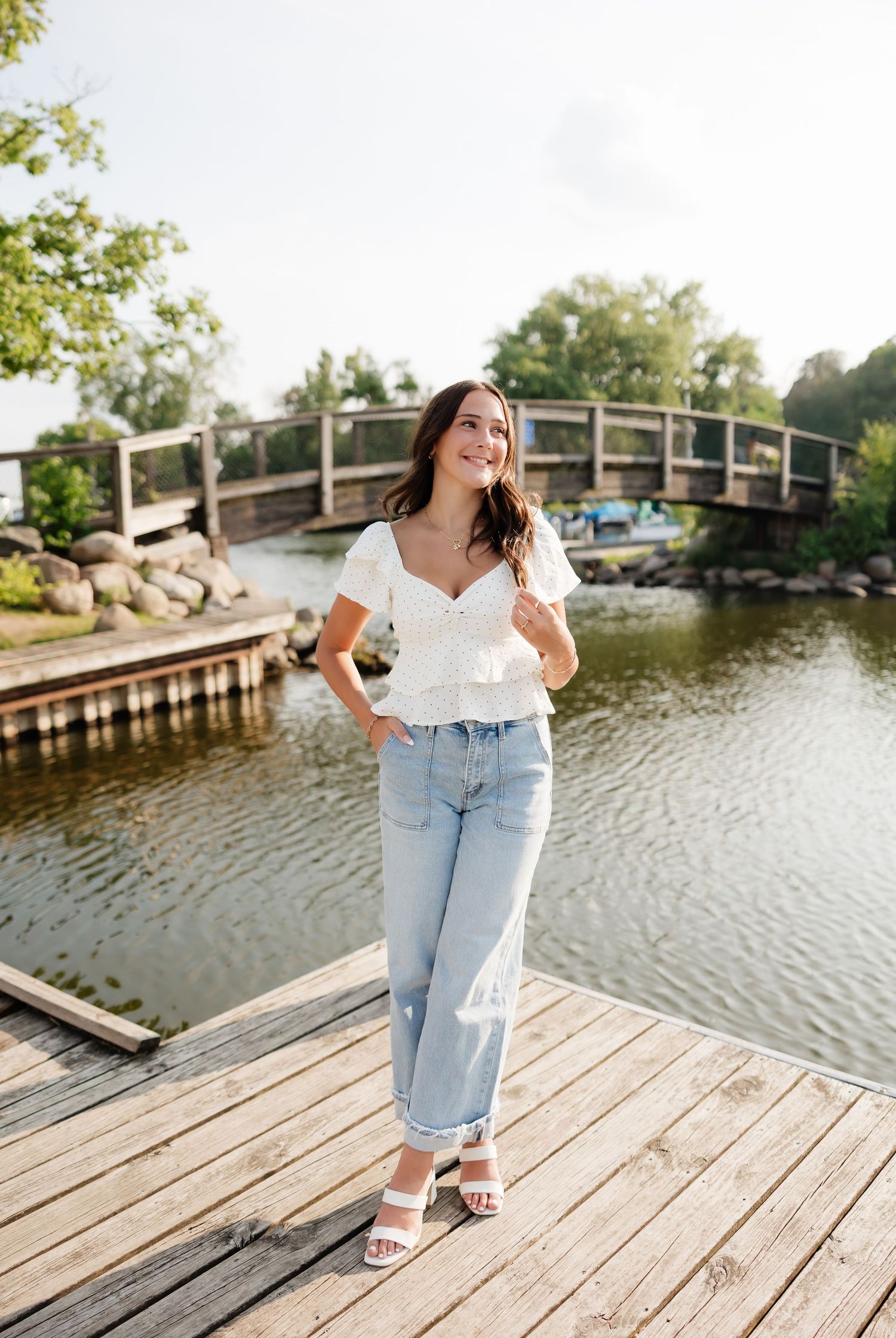 Young woman in white top and jeans poses on wooden dock by a serene waterway with a wooden bridge backdrop.