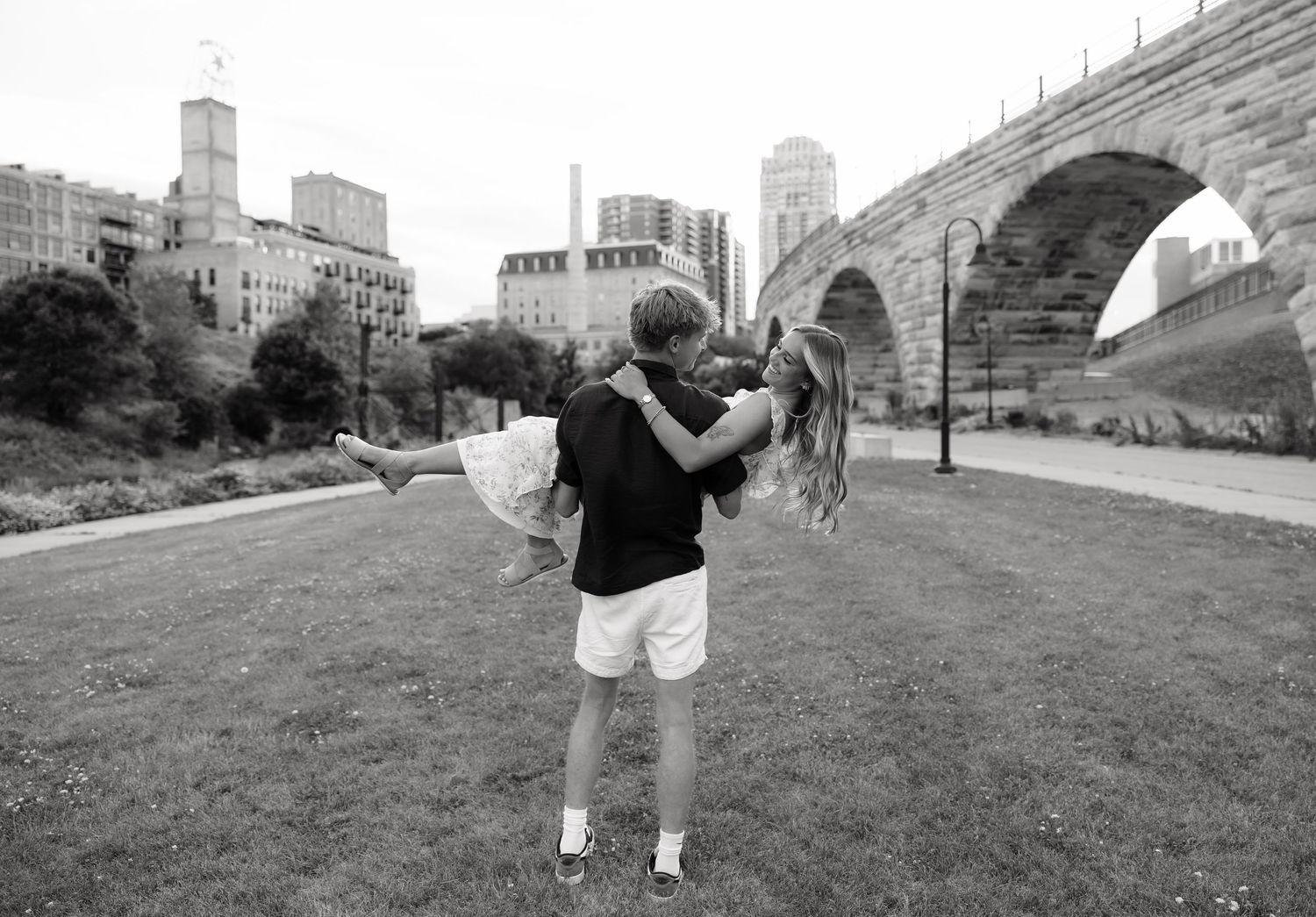 Black and white photo of a man lifting his partner in a romantic embrace near a stone bridge.