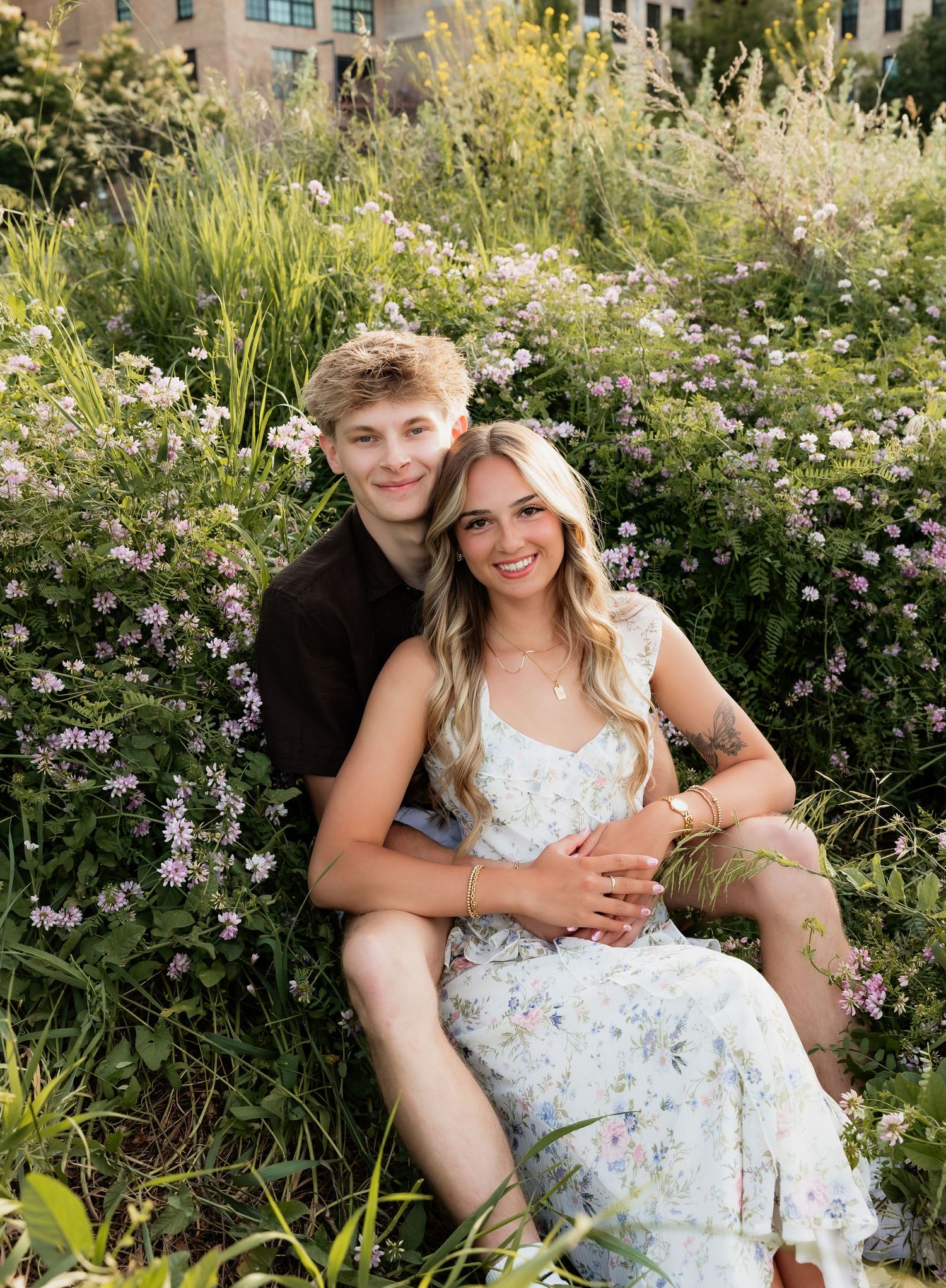 Couple sitting together in lush wildflower garden, smiling warmly, woman in floral dress, urban backdrop.