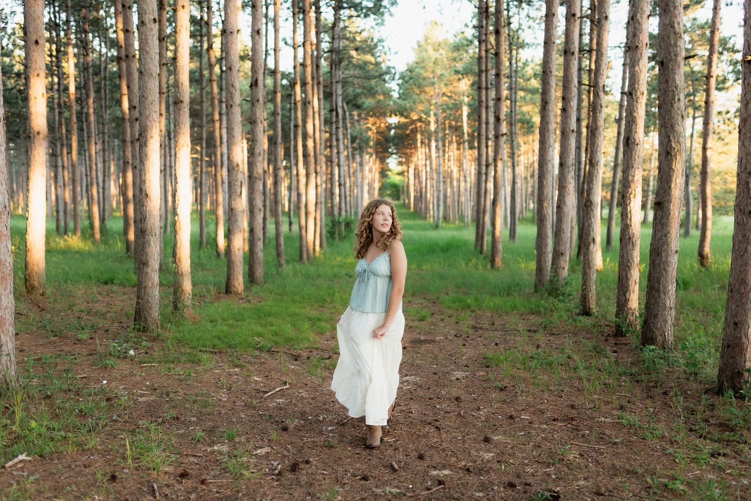 Woman in white dress walking through sunlit pine forest path smiling.