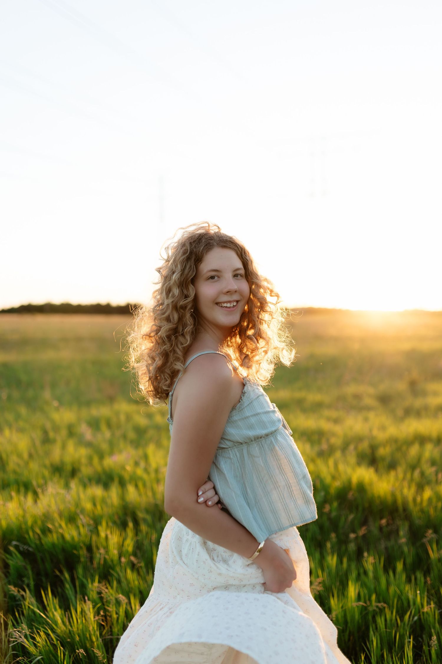 Young woman with curly hair smiling in a green field at golden sunset, wearing a blue top and white skirt.