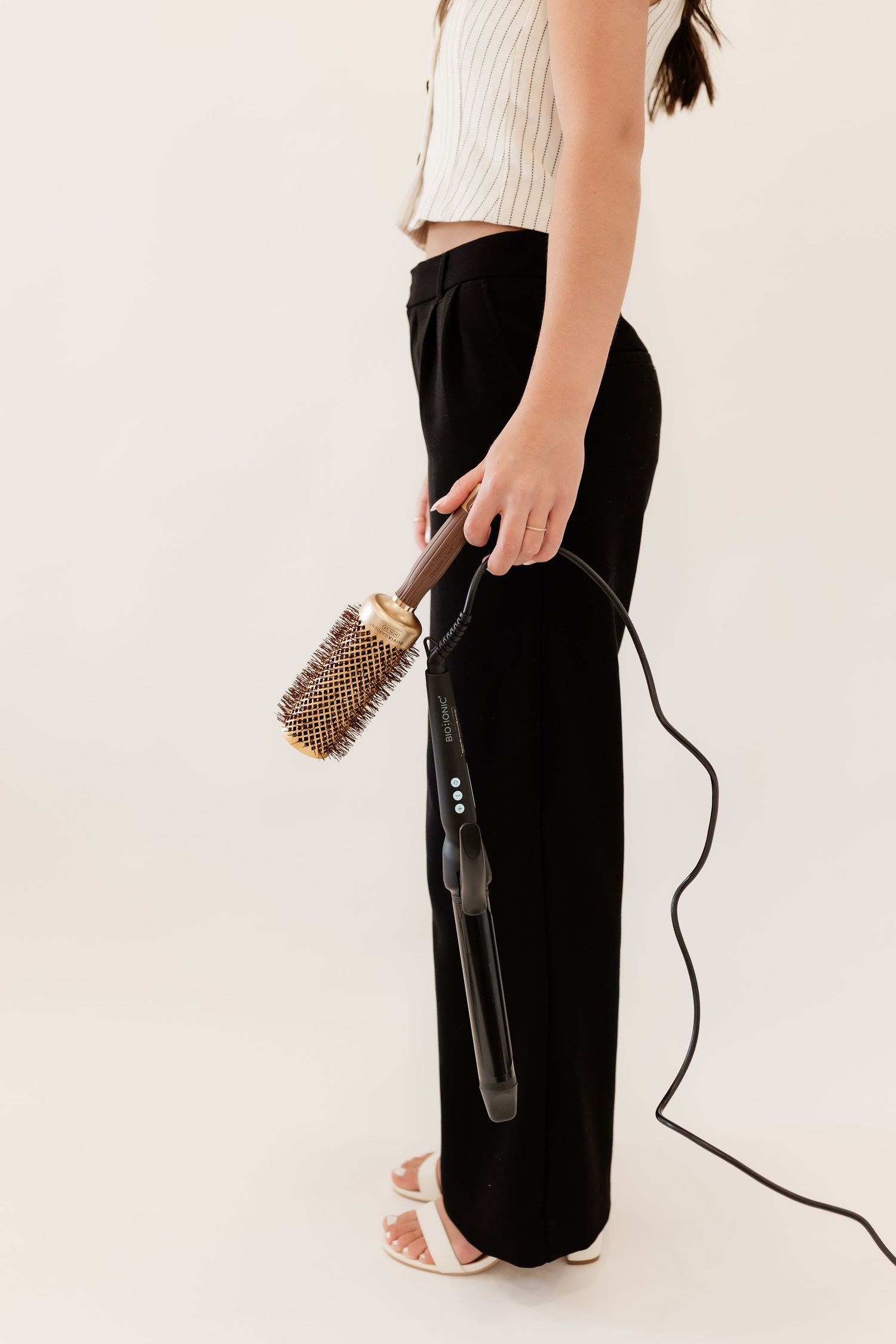 Woman in white crop top and black wide-leg pants holding a round brush and flat iron hair styling tools.