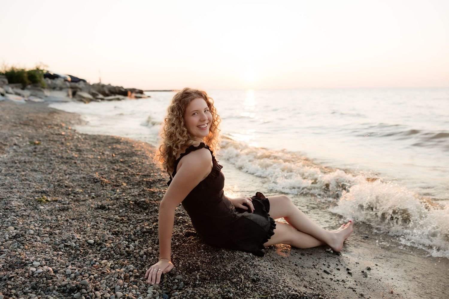 A smiling woman with curly hair sits on a pebble beach in a black dress at sunset.