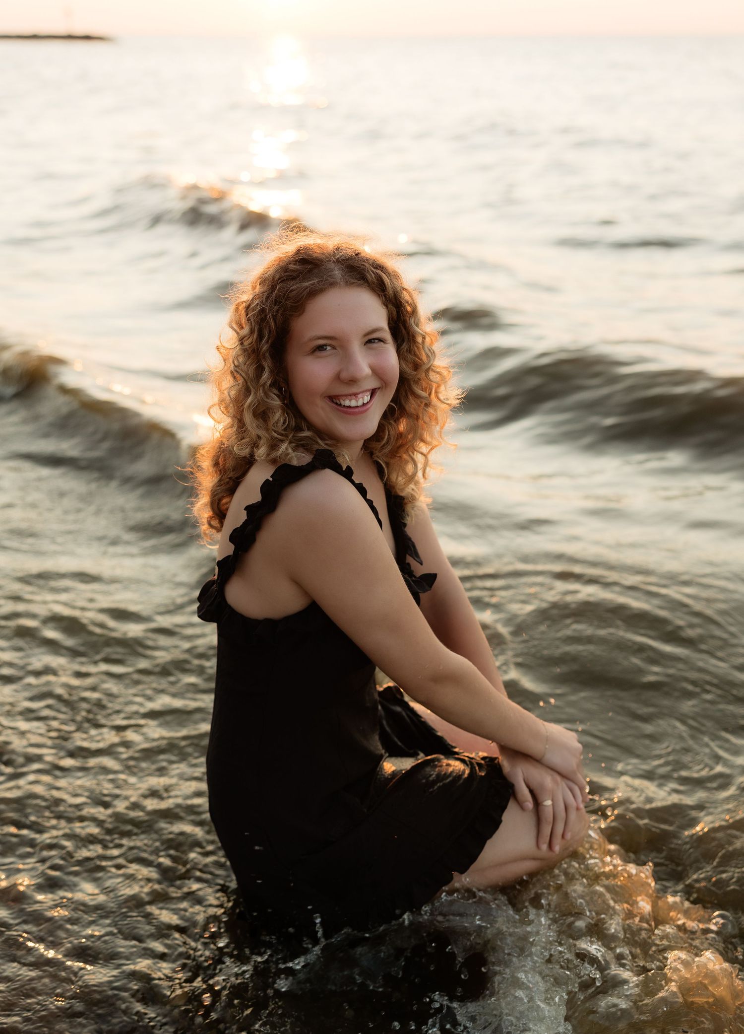 Woman with curly red hair smiling in ocean waves at golden hour sunset.