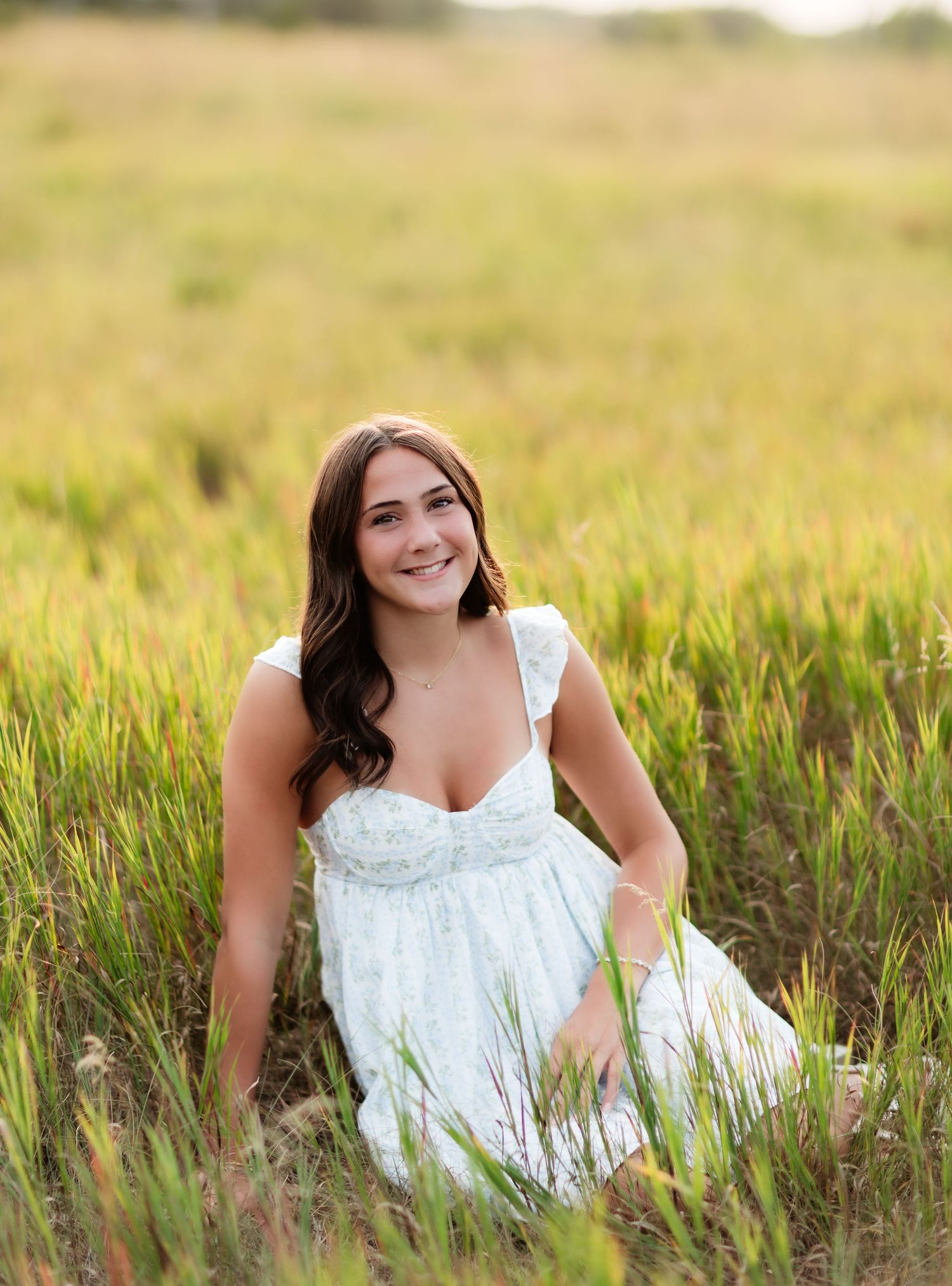 Brunette woman in white dress sitting in tall green grass field.