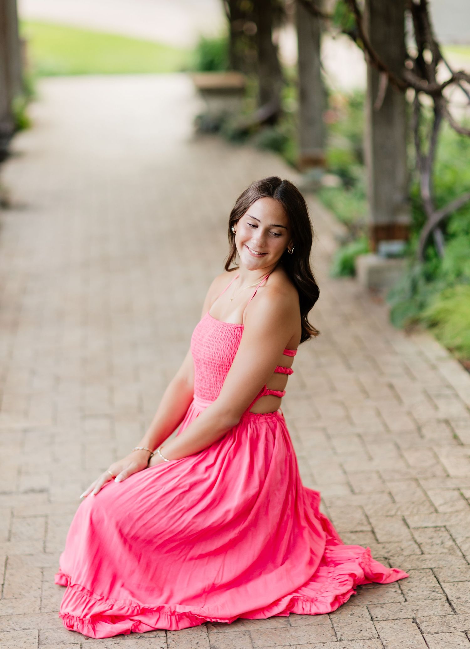 Brunette teen in hot pink halter maxi dress sitting gracefully on a brick garden pathway.