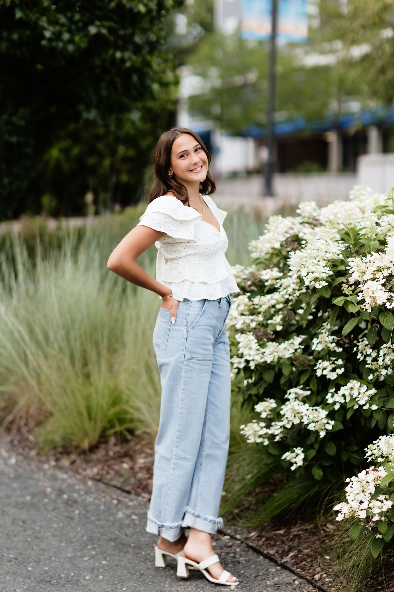Young woman in white ruffled top and blue wide-leg jeans posing beside white flowering bushes outdoors.