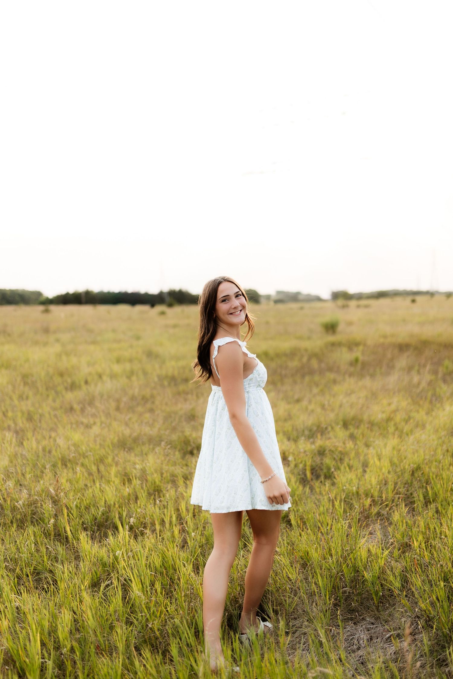 Smiling brunette woman in white dress standing in sunny meadow field.