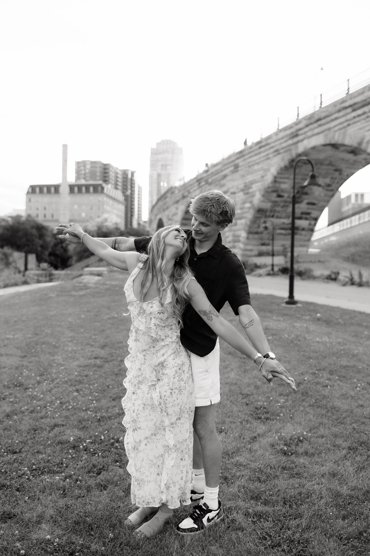 Black and white photo of a playful couple with outstretched arms near a stone arch bridge in an urban park.