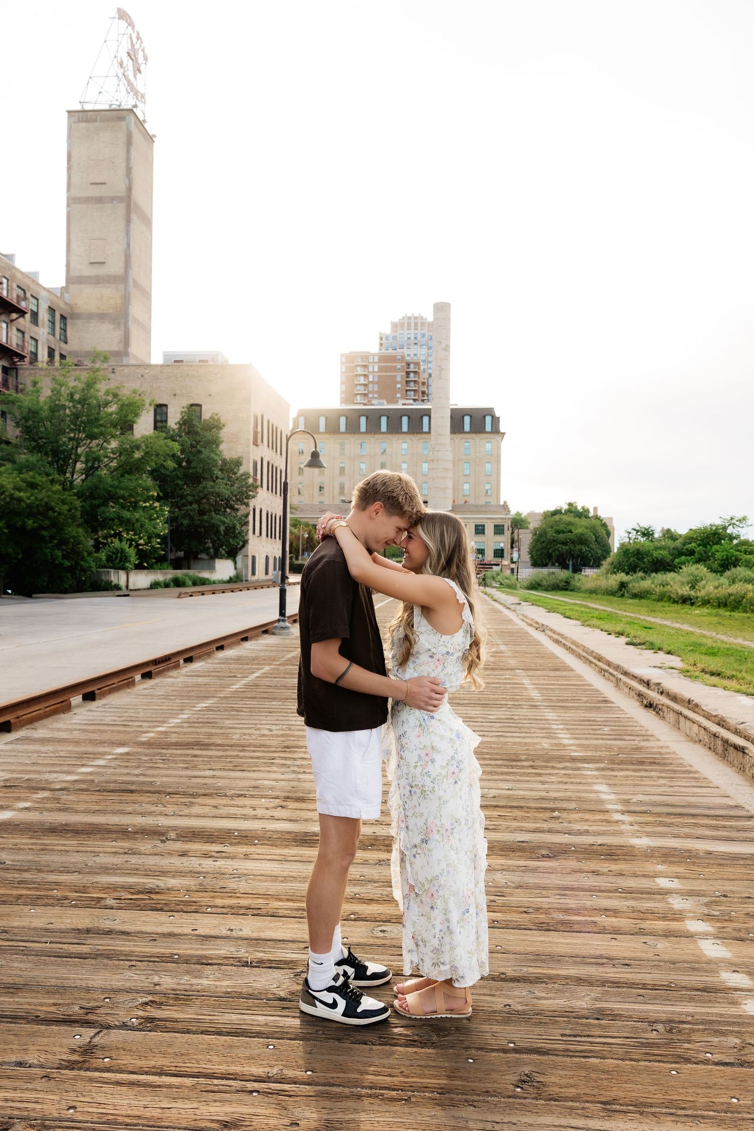 Romantic couple sharing a tender moment, touching foreheads on a wooden boardwalk with city buildings in background.