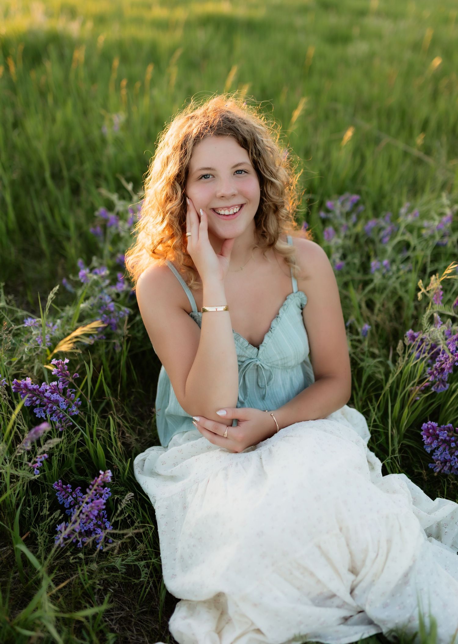 Joyful young woman with curly hair sitting among purple wildflowers in a green meadow at sunset.