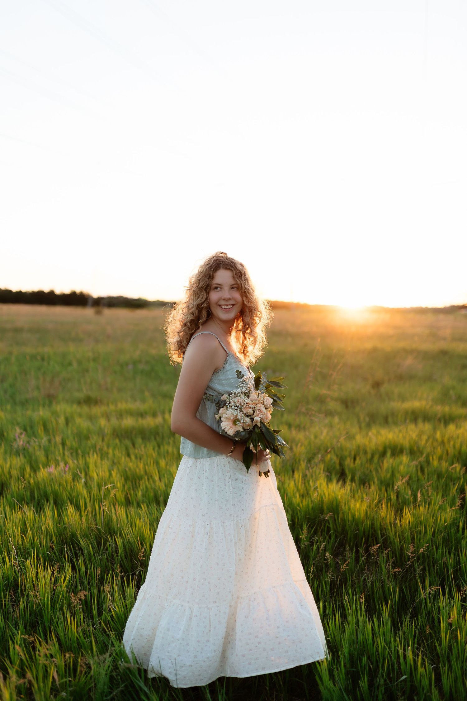 Smiling curly-haired woman in white tiered dress holding flowers in a sunlit green field at sunset.