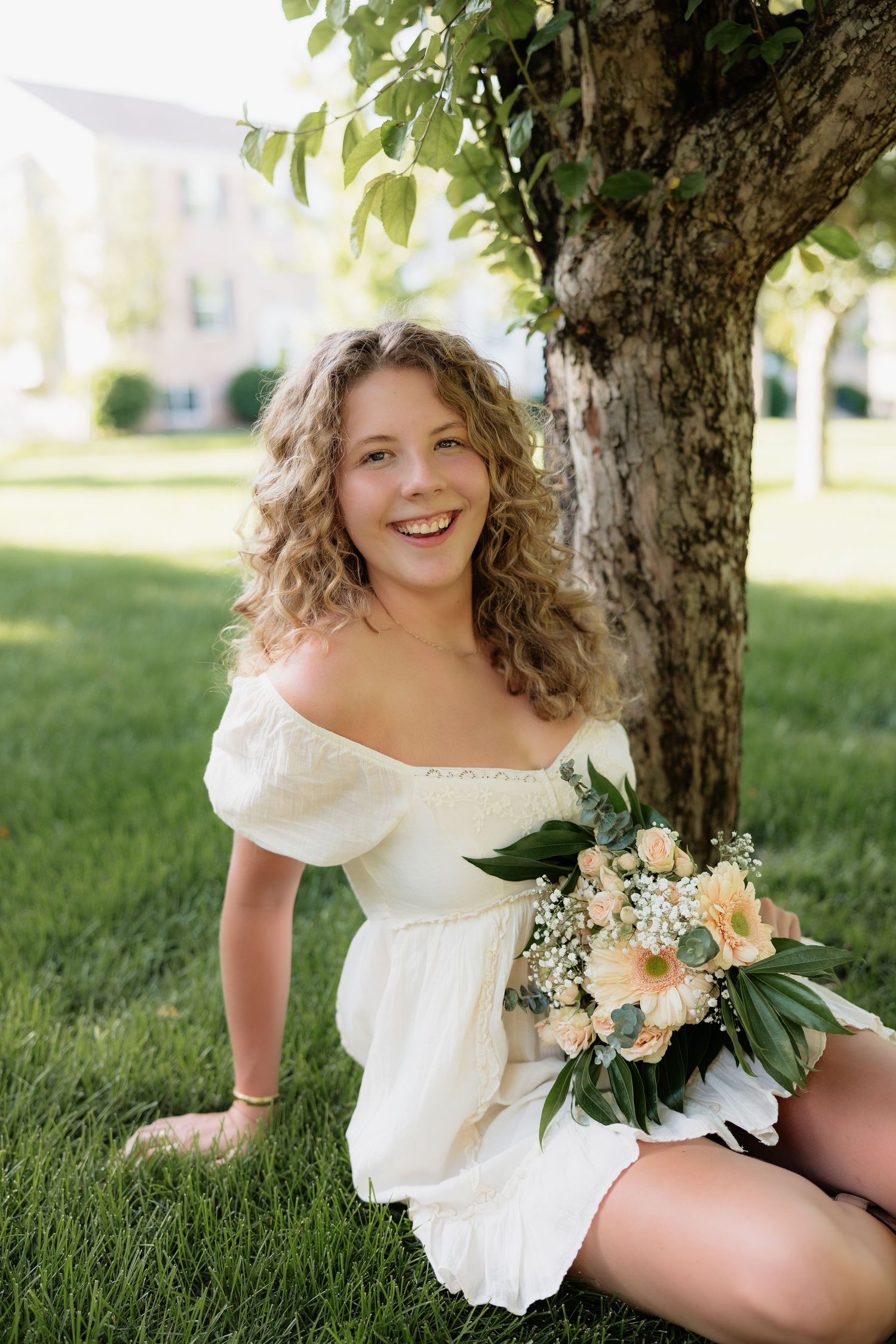 Smiling young woman in white off-shoulder dress holding a bouquet of flowers while sitting on grass under a tree.
