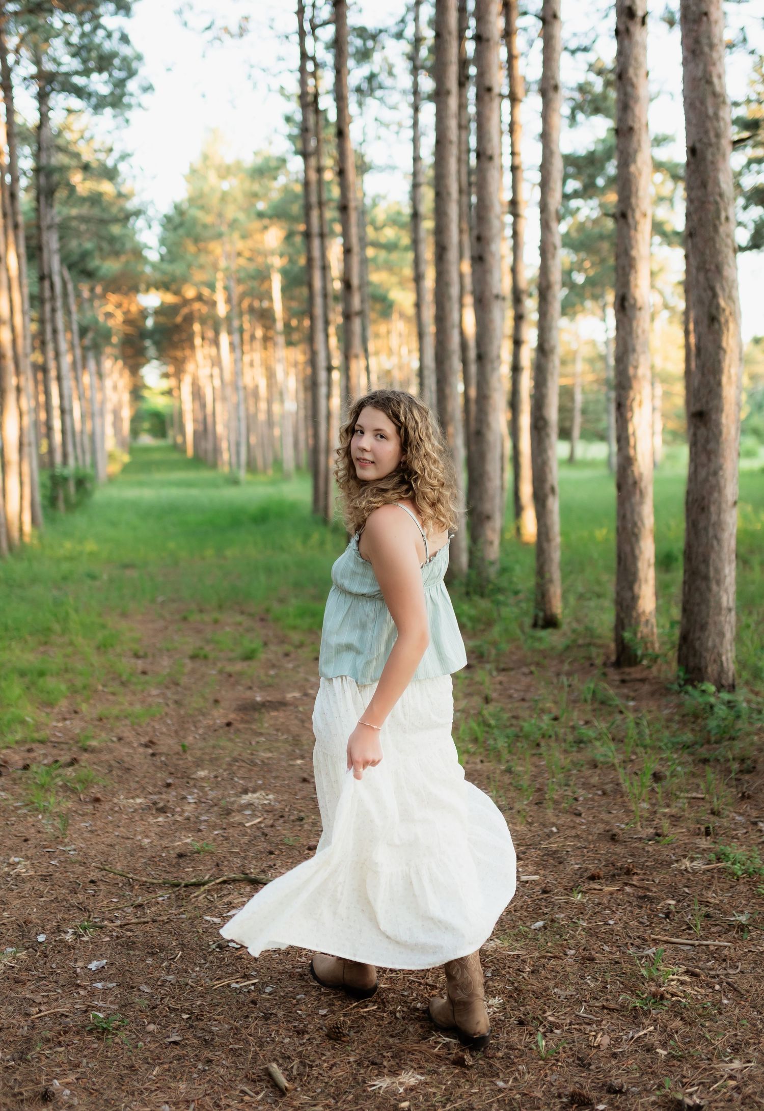 Young woman in mint top and white skirt twirls among tall pine trees in golden hour light.