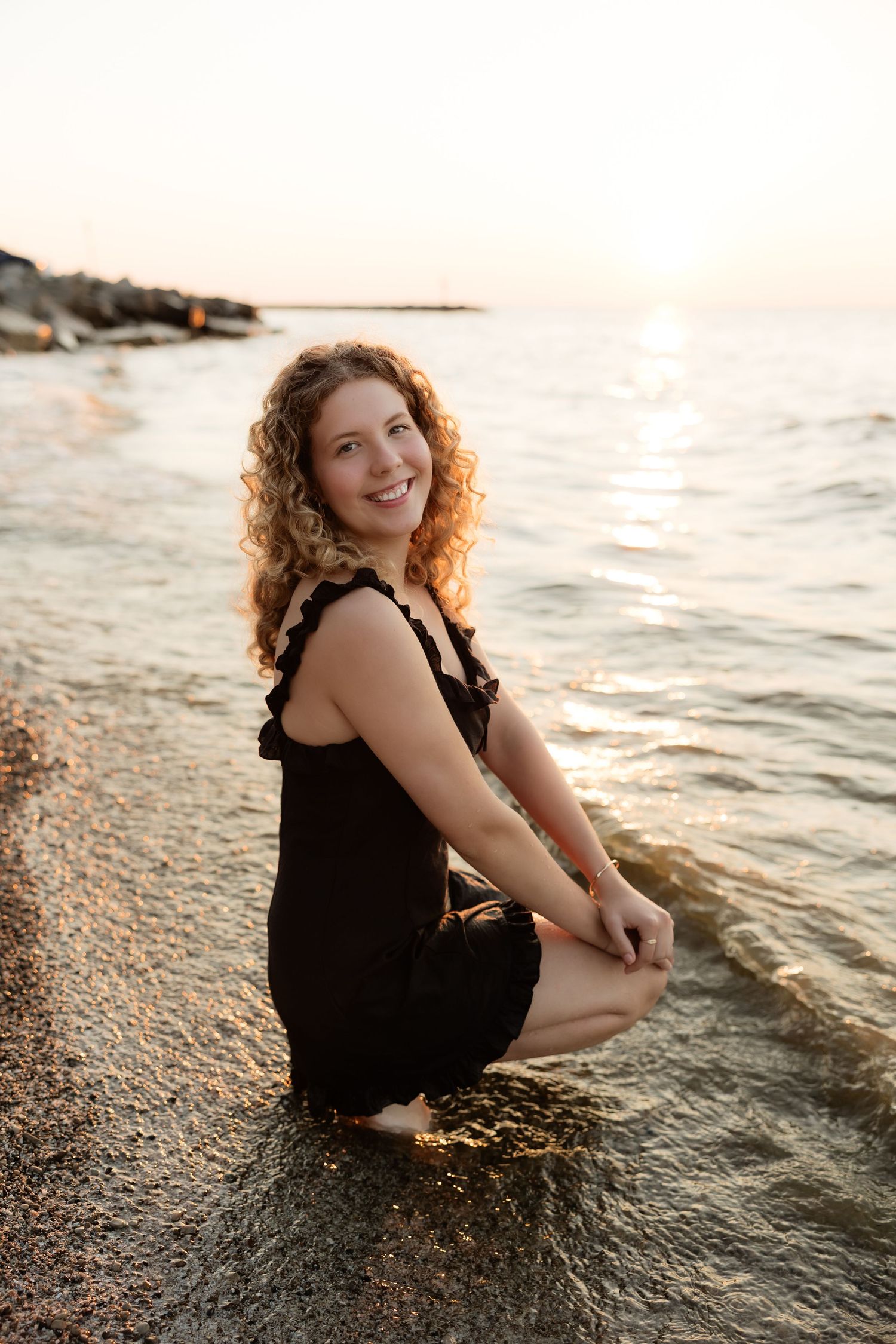 Young woman with curly hair in black dress sitting at water's edge on rocky beach at sunset.