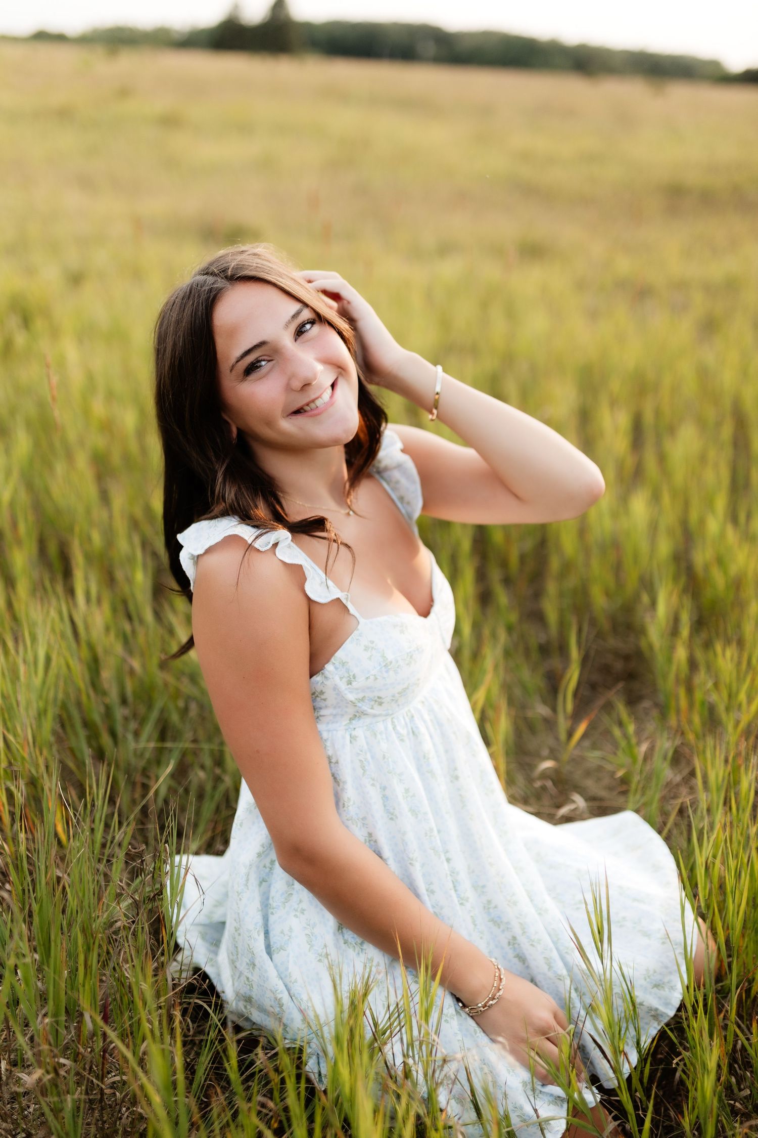 Smiling young woman in a white sundress sits in a golden grass field, brushing hair from her face.