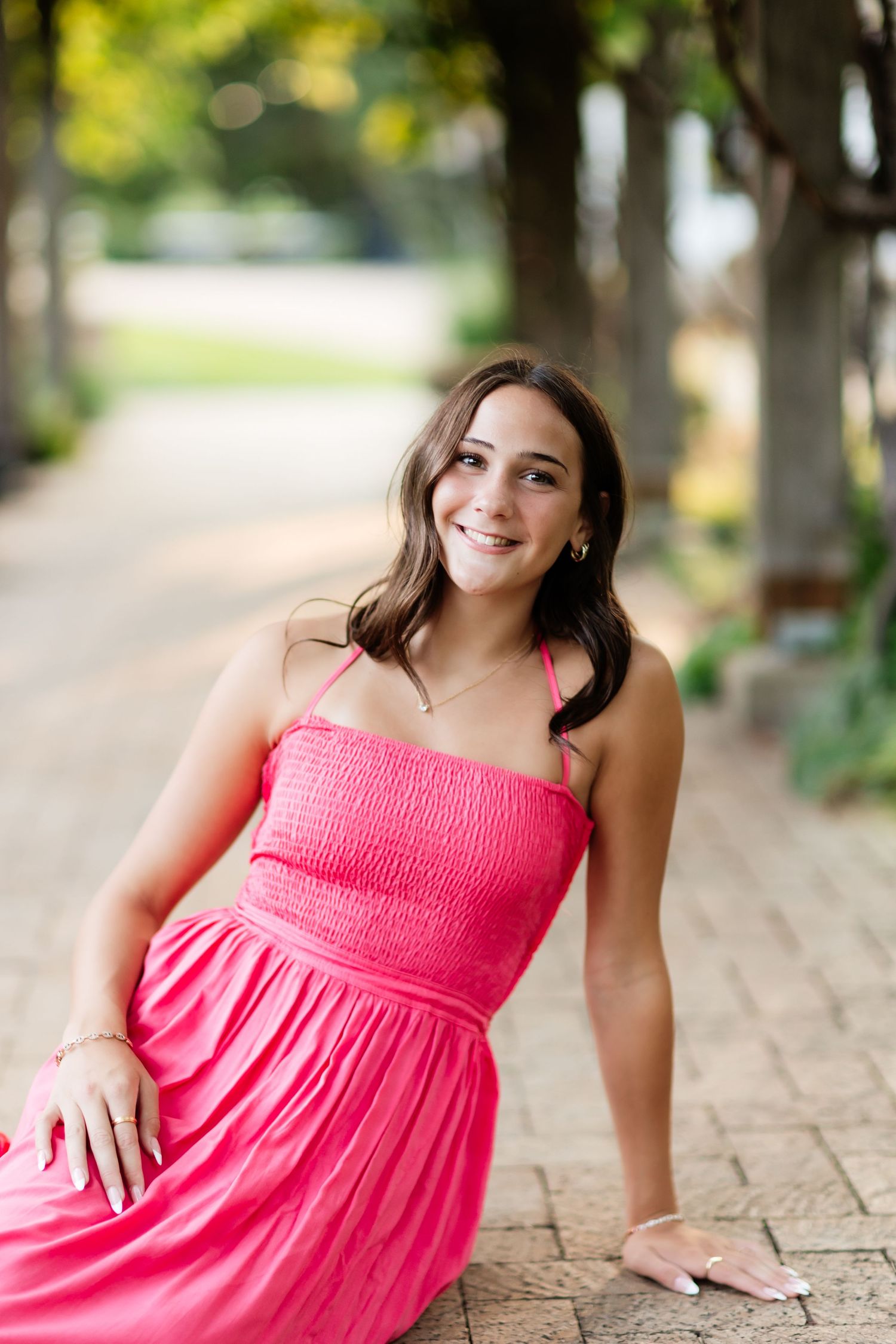 Smiling young woman in pink smocked maxi dress sitting on a brick pathway surrounded by trees.