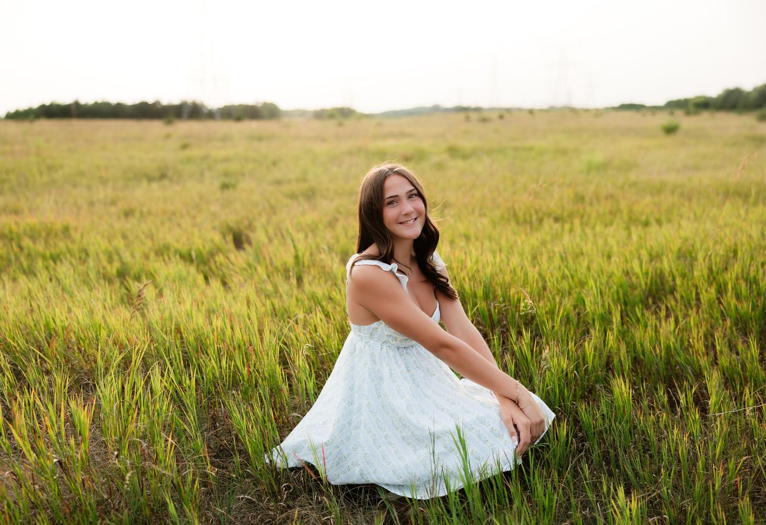 Young woman in a white sundress sits gracefully in a golden green meadow field on a cloudy day.