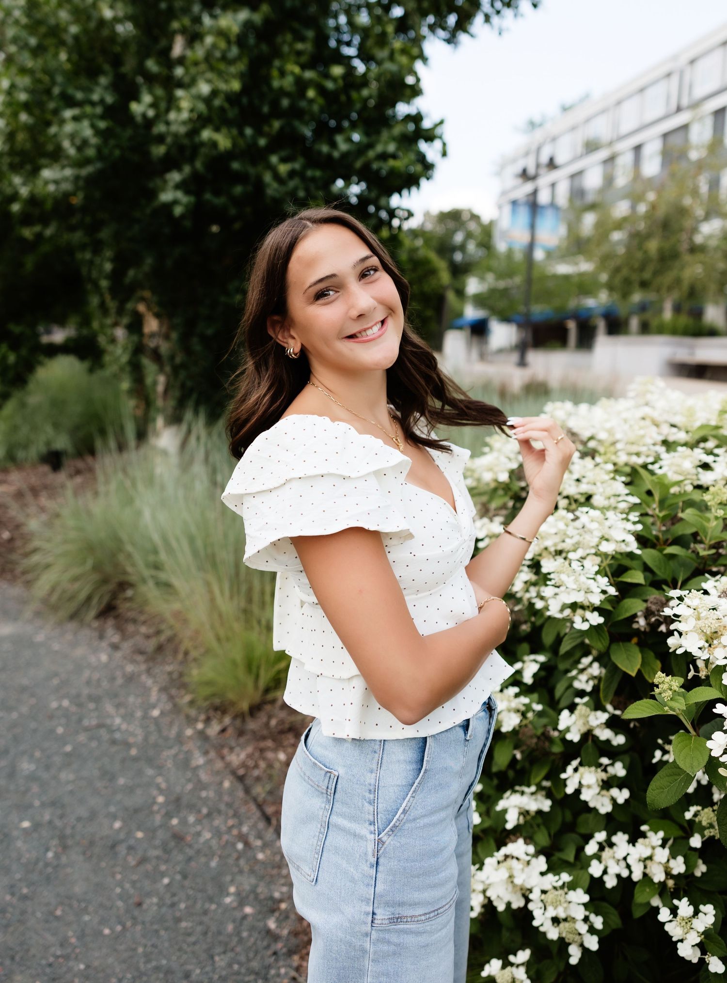 Smiling brunette woman in white floral top and jeans posing next to white hydrangea flowers outside.
