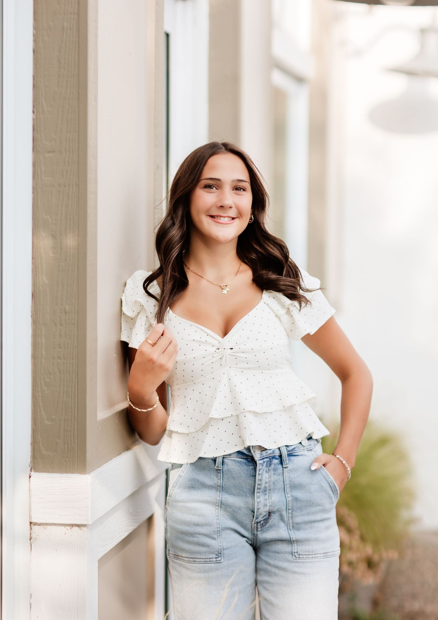 Young woman in white ruffle top and light jeans posing against white column exterior wall.
