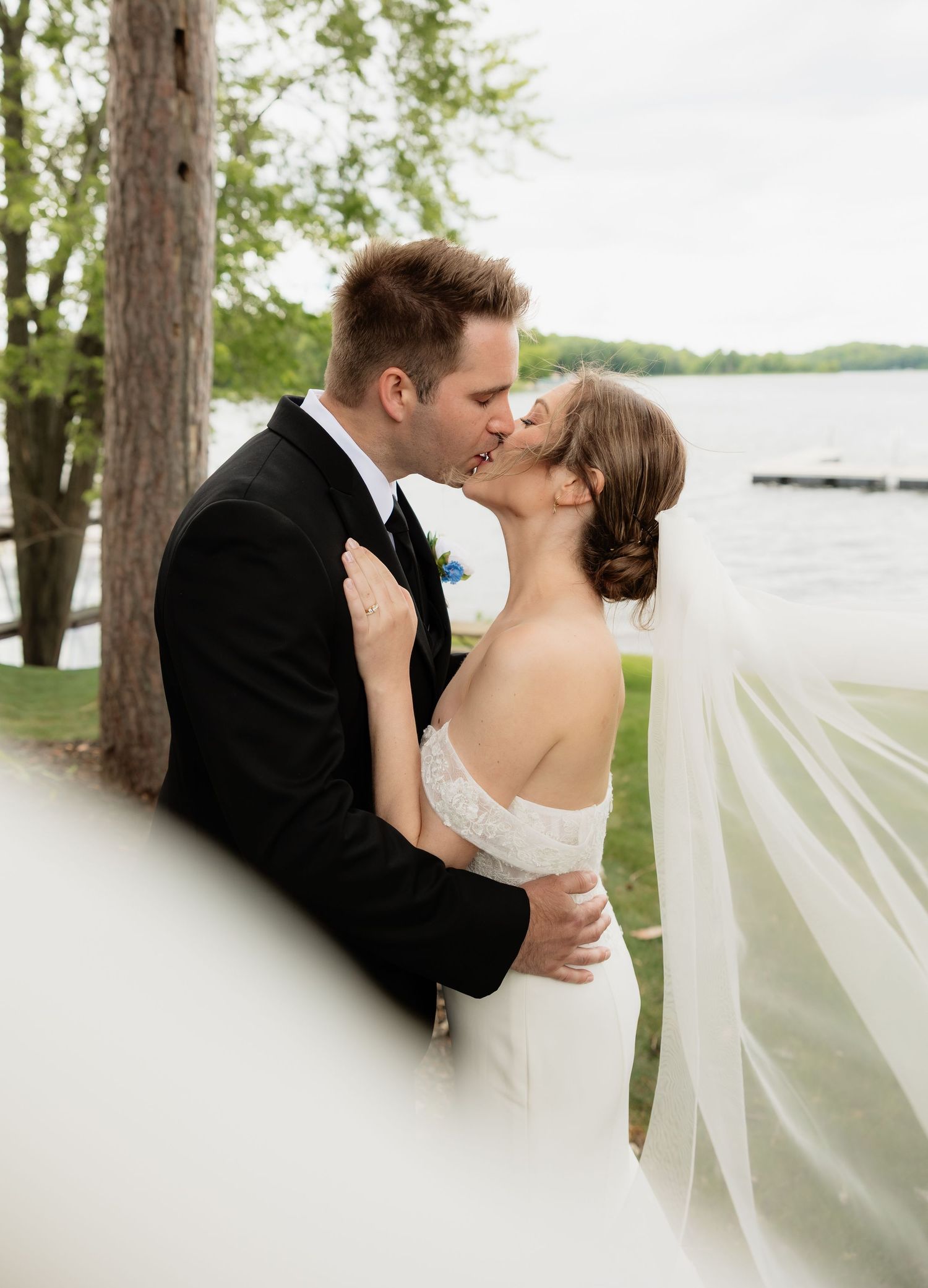 Bride and groom share a romantic kiss outdoors near a lake, with her white veil flowing in the breeze.