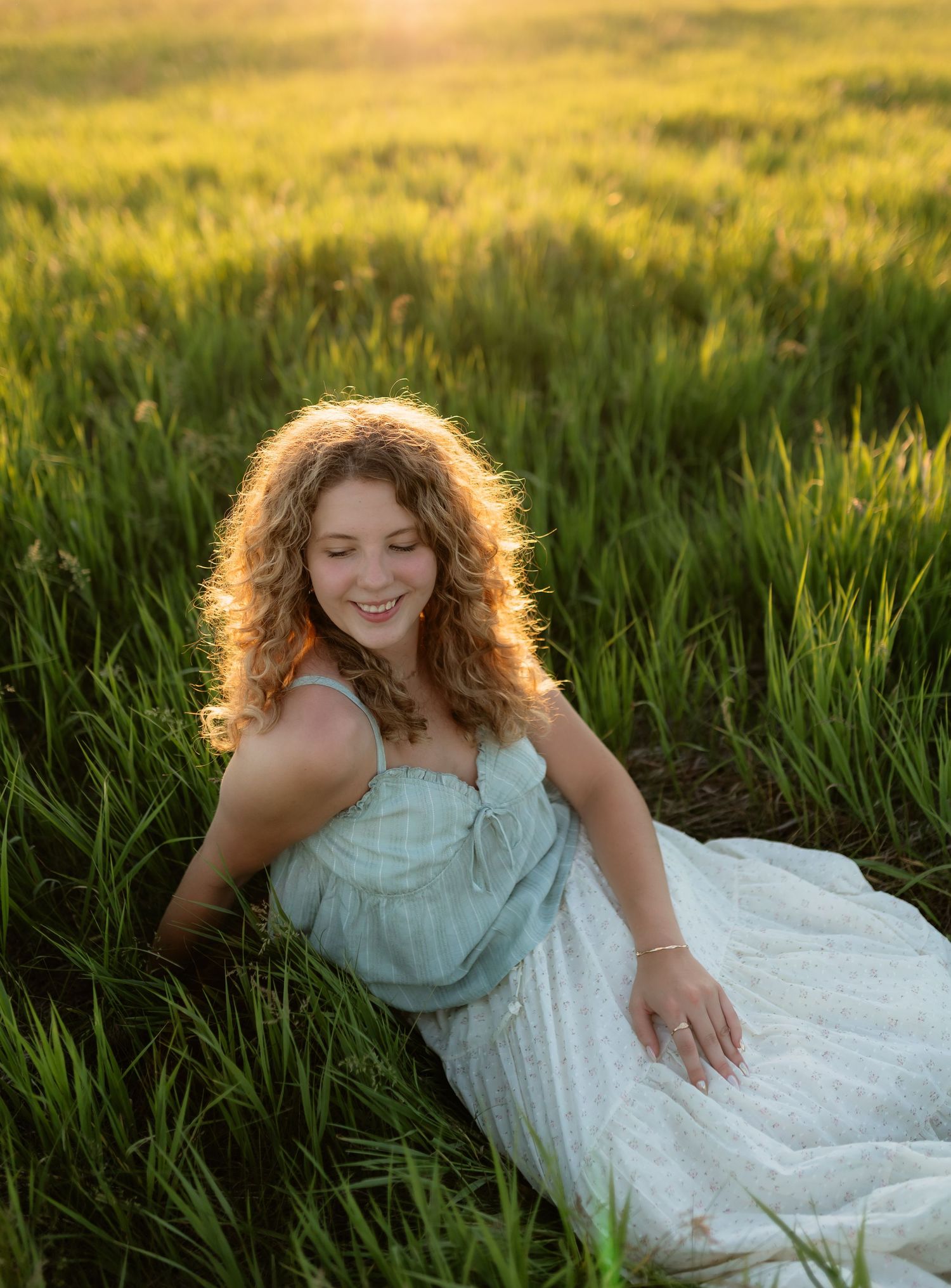 Young woman with curly hair smiling in a green field at golden hour, wearing a light blue dress.