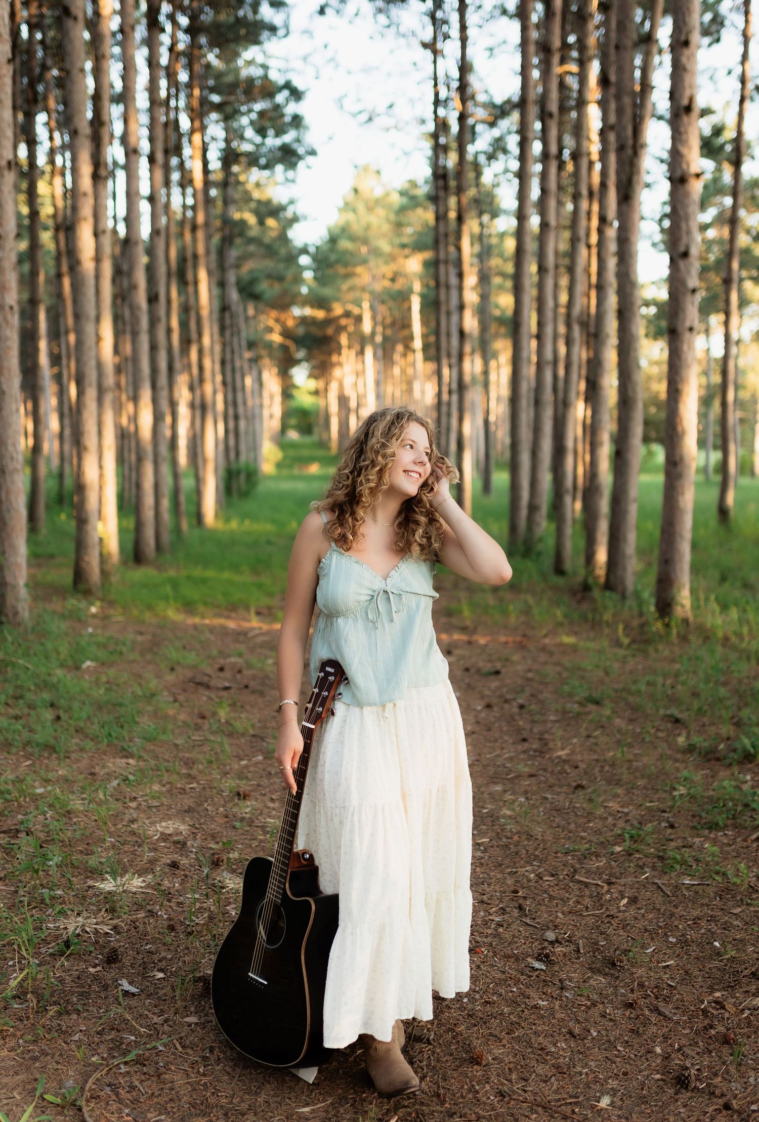Young woman in mint top and white skirt holding a guitar while standing among tall pine trees.